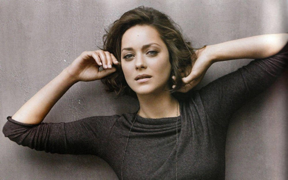 Actress HD 桌布 壁紙 — 一位擁有波浪形棕色頭髮的女性靠著紋理灰色牆壁，雙手輕輕握住頭髮，穿著深灰色毛衣。