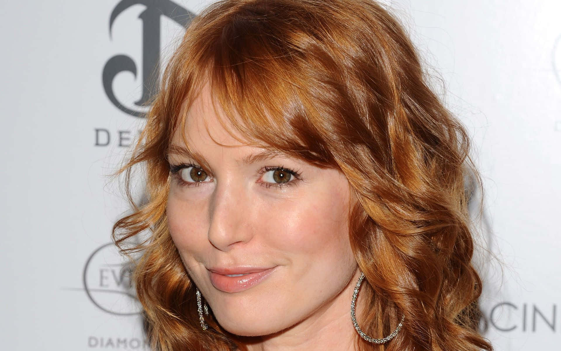 Alicia Witt 桌布 壁紙 — 一位擁有波浪形赤褐色頭髮和銀色圓環耳環的女性近距離肖像，對著鏡頭溫柔微笑。