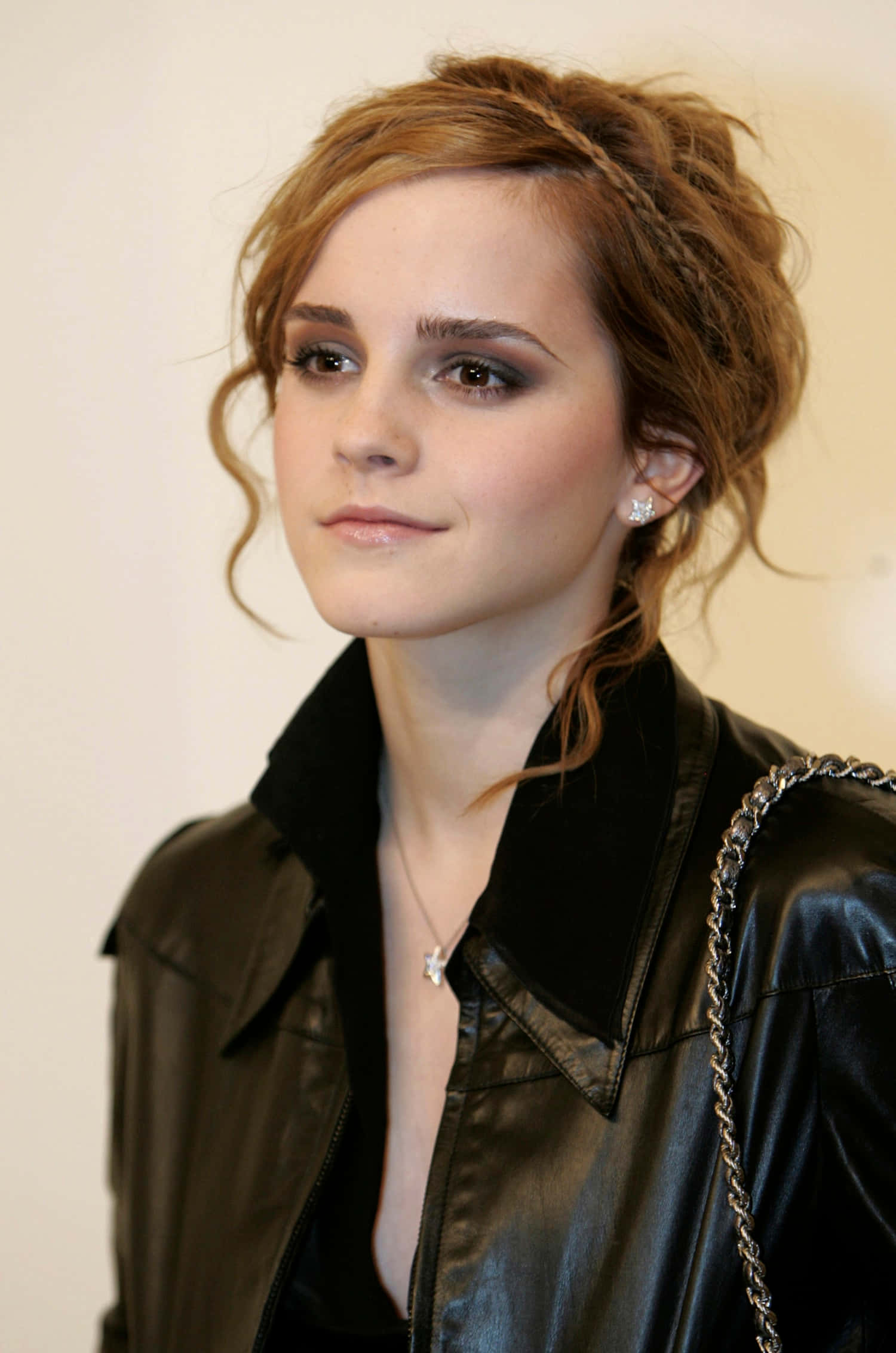 Emma Watson 桌布 壁紙 — 一位擁有波浪形赤褐色頭髮的女性，穿著黑色皮夾克和鑽石耳環，背景是純色米色牆壁。