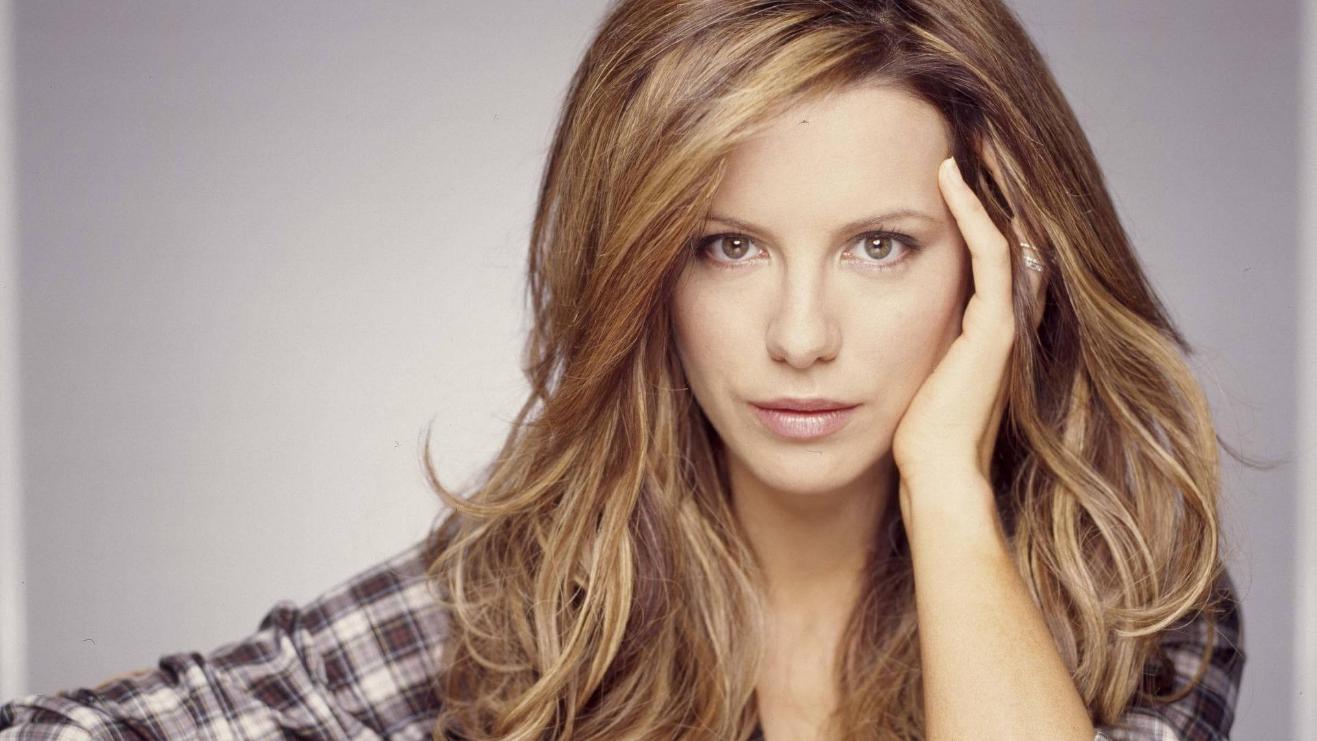 Kate Beckinsale 桌布 壁紙 — 一位擁有波浪形金髮的女性，在純灰色背景下穿著格紋襯衫，將頭靠在手上。