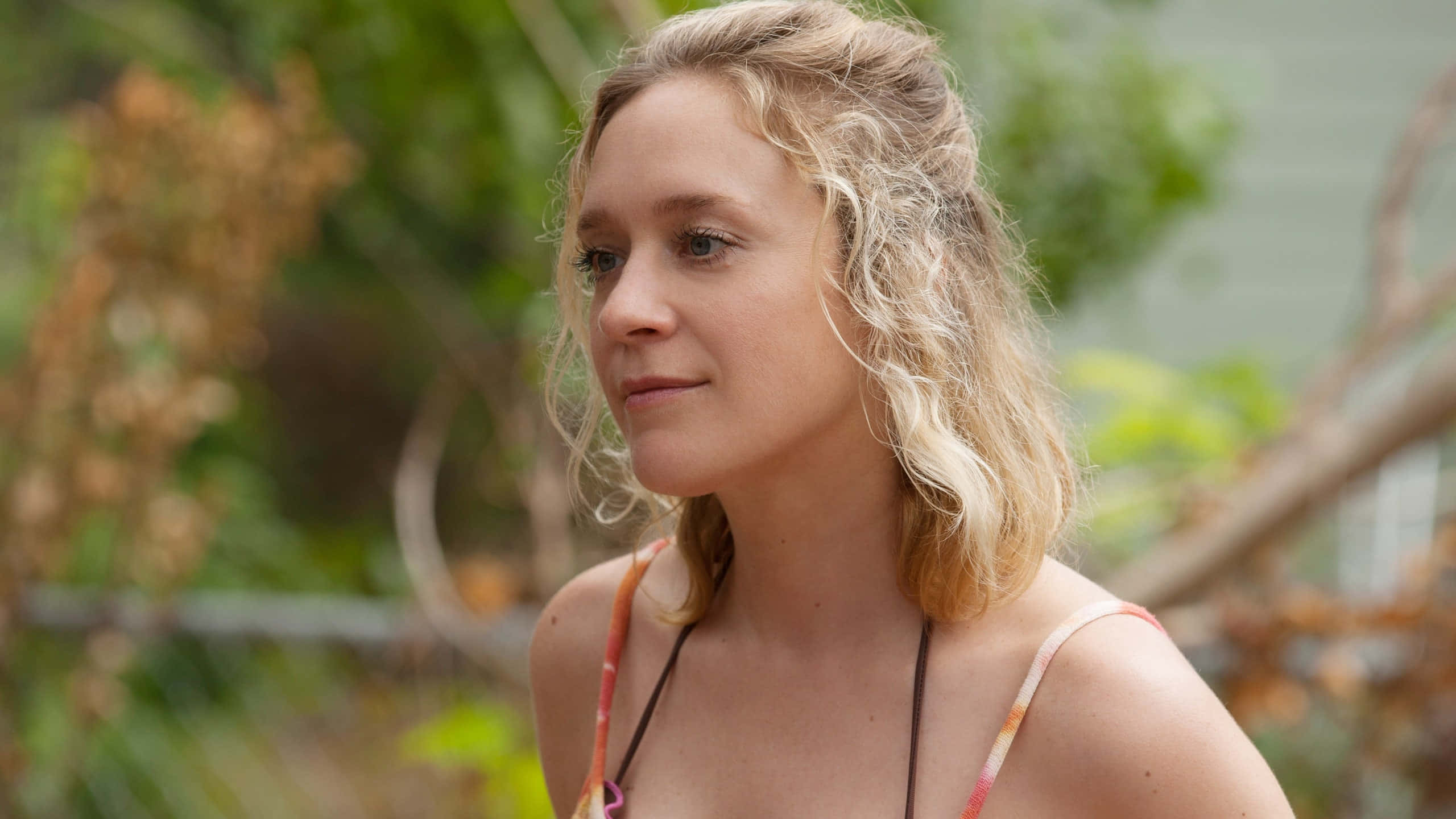 Chloë Sevigny 桌布 壁紙 — 一位擁有波浪形金髮的女性在戶外，在茂密的綠色樹葉和柔和自然光線中若有所思地凝視。
