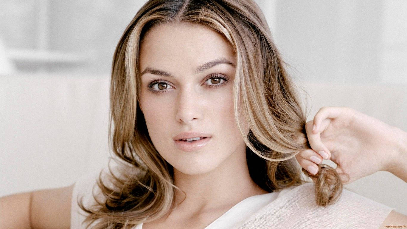 Keira Knightley 桌布 壁紙 — 一位擁有波浪形金髮的女性在柔和光線的攝影棚中，輕輕拉動一縷髮絲，同時直視鏡頭。