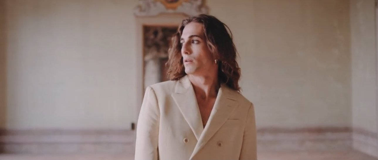 Damiano David 桌布 壁紙 — 一位擁有波浪形頭髮、身穿奶油色西裝外套的人，站在一個宏偉、光線柔和且裝飾華麗的房間裡。