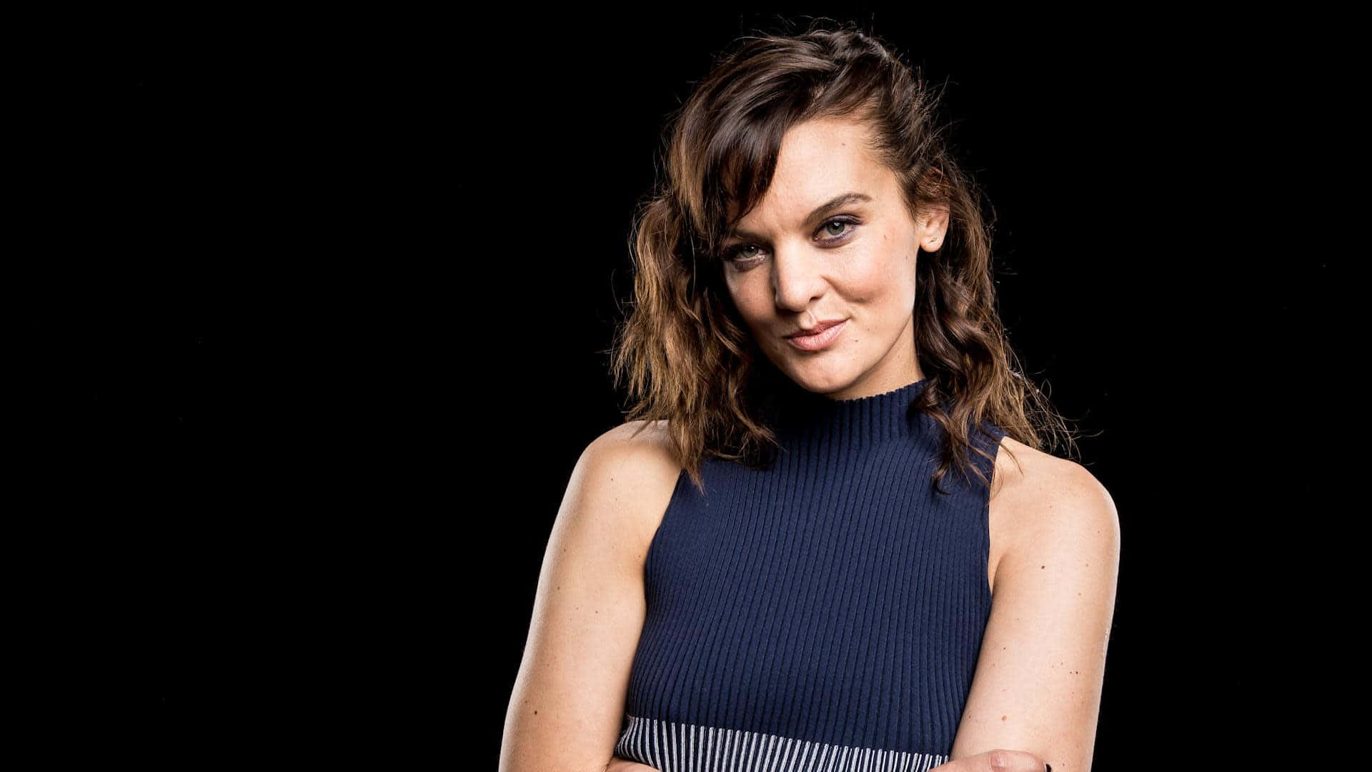 Frankie Shaw 桌布 壁紙 — 一位擁有波浪棕色長髮、身穿海軍藍羅紋上衣的女性，在純黑背景前自信地擺姿勢。