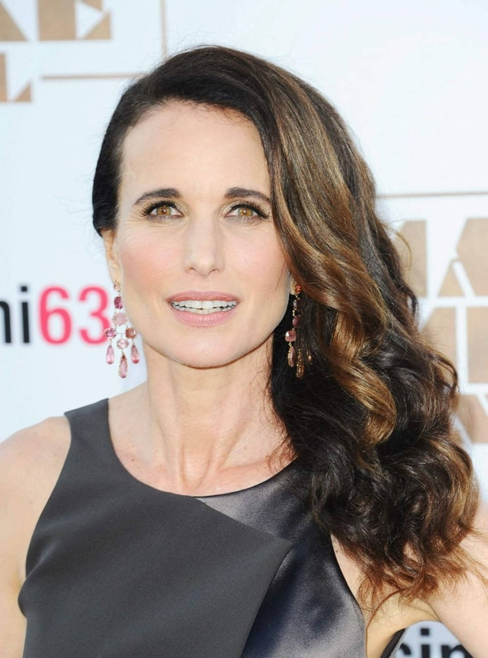 andie macdowell 桌布 壁紙 — 一位擁有波浪棕色長髮和優雅垂墜耳環的女性，在活動中穿著時尚黑色連衣裙對著鏡頭微笑。