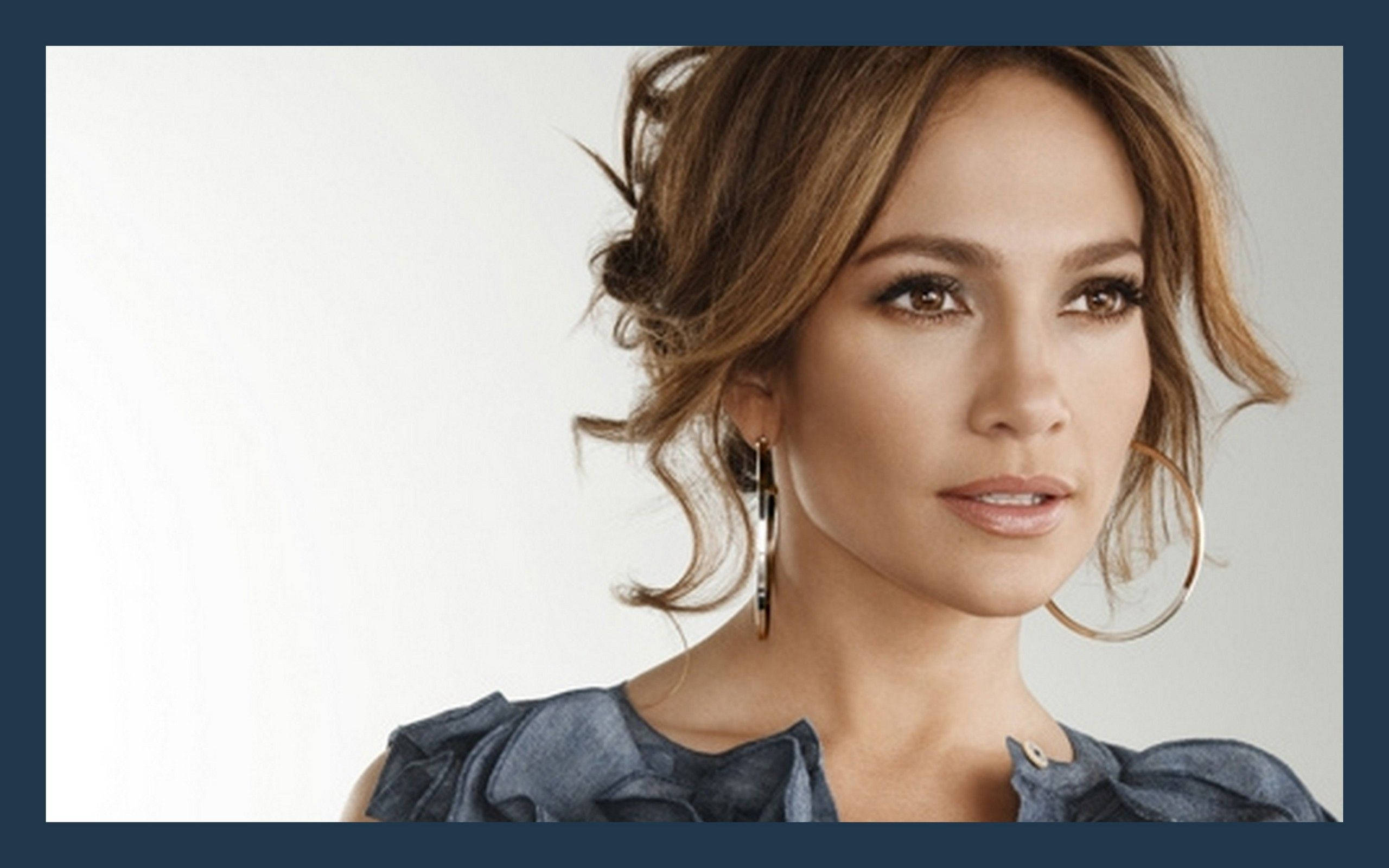 Jennifer Lopez 桌布 壁紙 — 一位擁有波浪棕色長髮和大圈耳環的女性，穿著藍色荷葉邊上衣，以純白色背景為襯的近距離肖像。