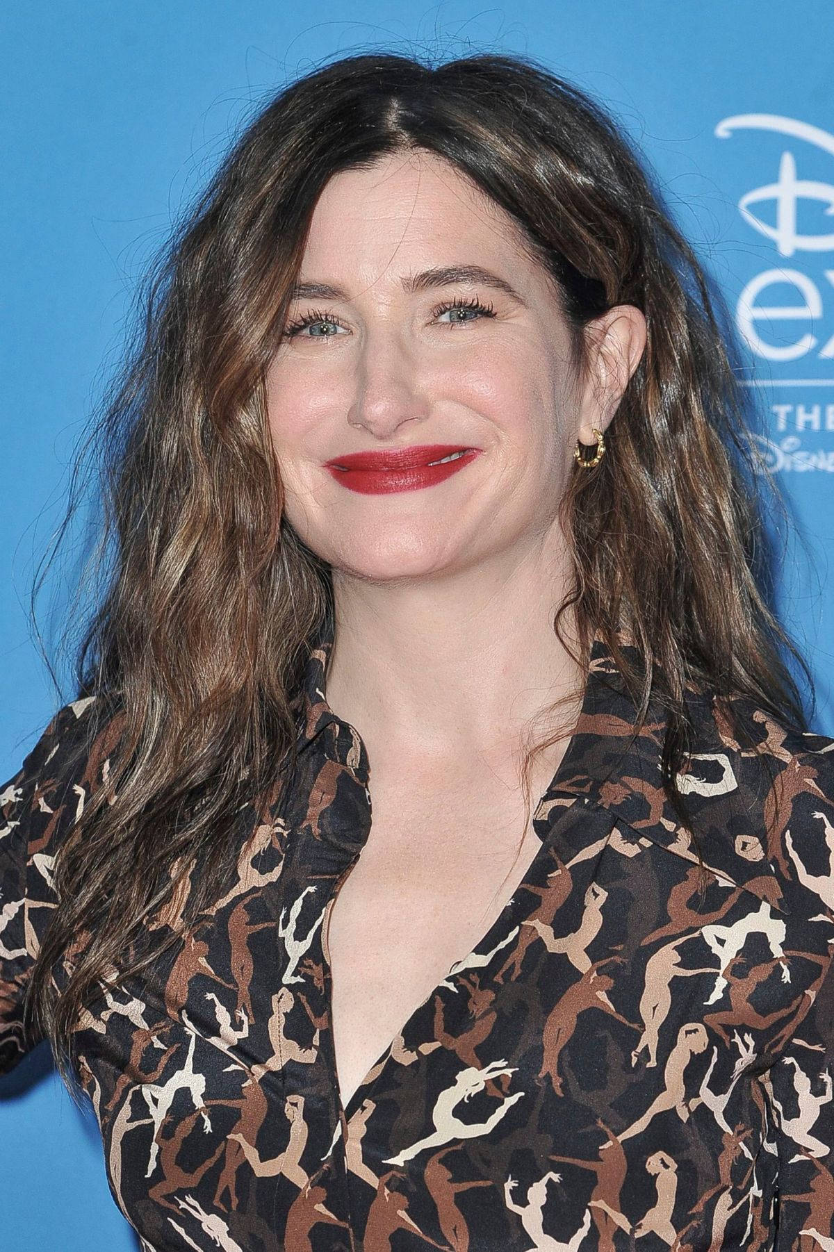 Celebrity 桌布 壁紙 — 一位擁有波浪棕色長髮和紅色唇膏的女性，在藍色迪士尼活動背景前溫暖微笑。