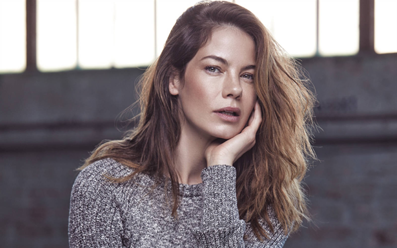 Michelle Monaghan 桌布 壁紙 — 一位擁有波浪棕色長髮的女性，將下巴擱在手上，靠在一面帶有大窗戶的磚牆前。