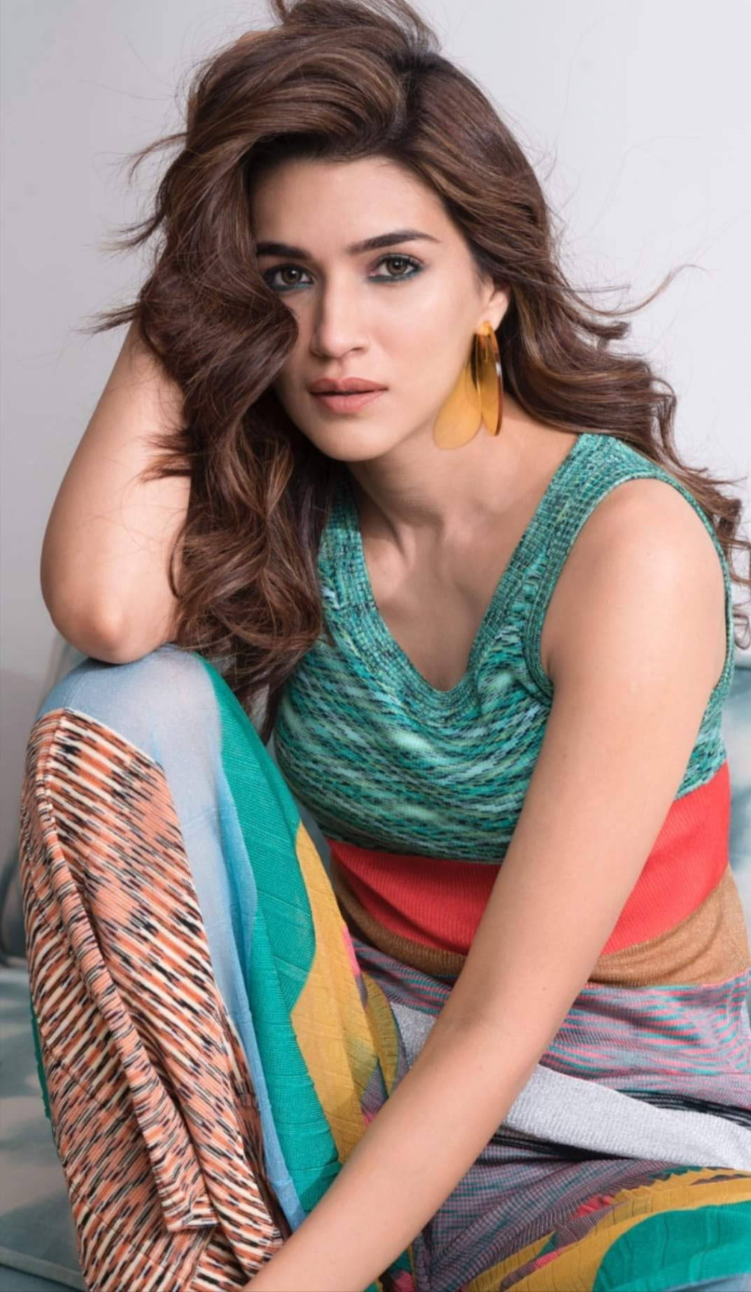 Actress HD 桌布 壁紙 — 一位擁有波浪棕色長髮的女性，穿著色彩繽紛的圖案服裝，在純色背景前自信地擺姿勢。