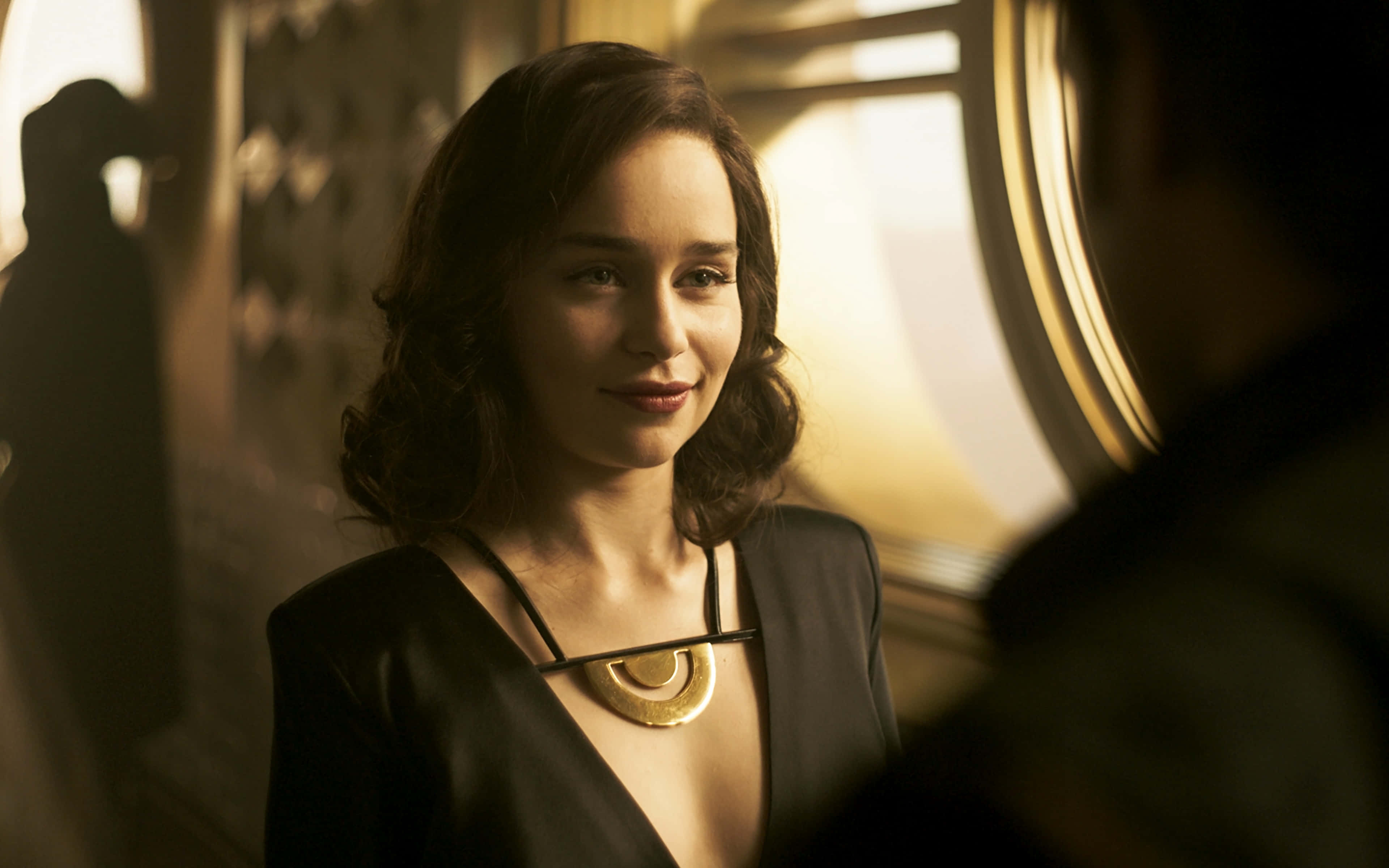 Solo A Star Wars Story 桌布 壁紙 — 一位擁有波浪棕色長髮的女性，穿著黑色連衣裙和金色項鍊，在陽光灑落的火車車廂內溫暖地微笑。