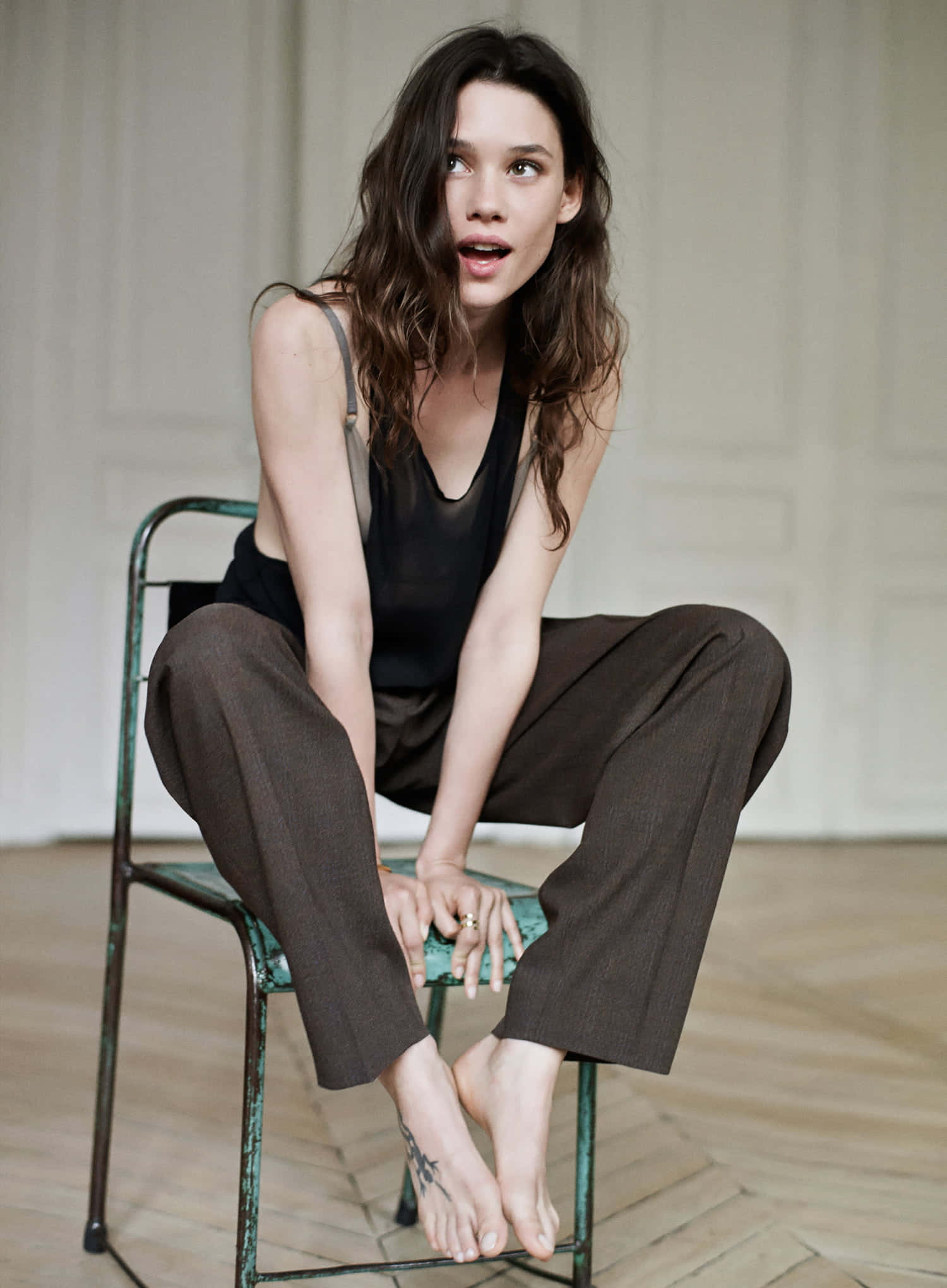 Astrid Berges Frisbey 桌布 壁紙 — 一位擁有波浪棕色長髮的女性赤腳坐在復古綠色椅子上，房間簡約，鋪有木地板。