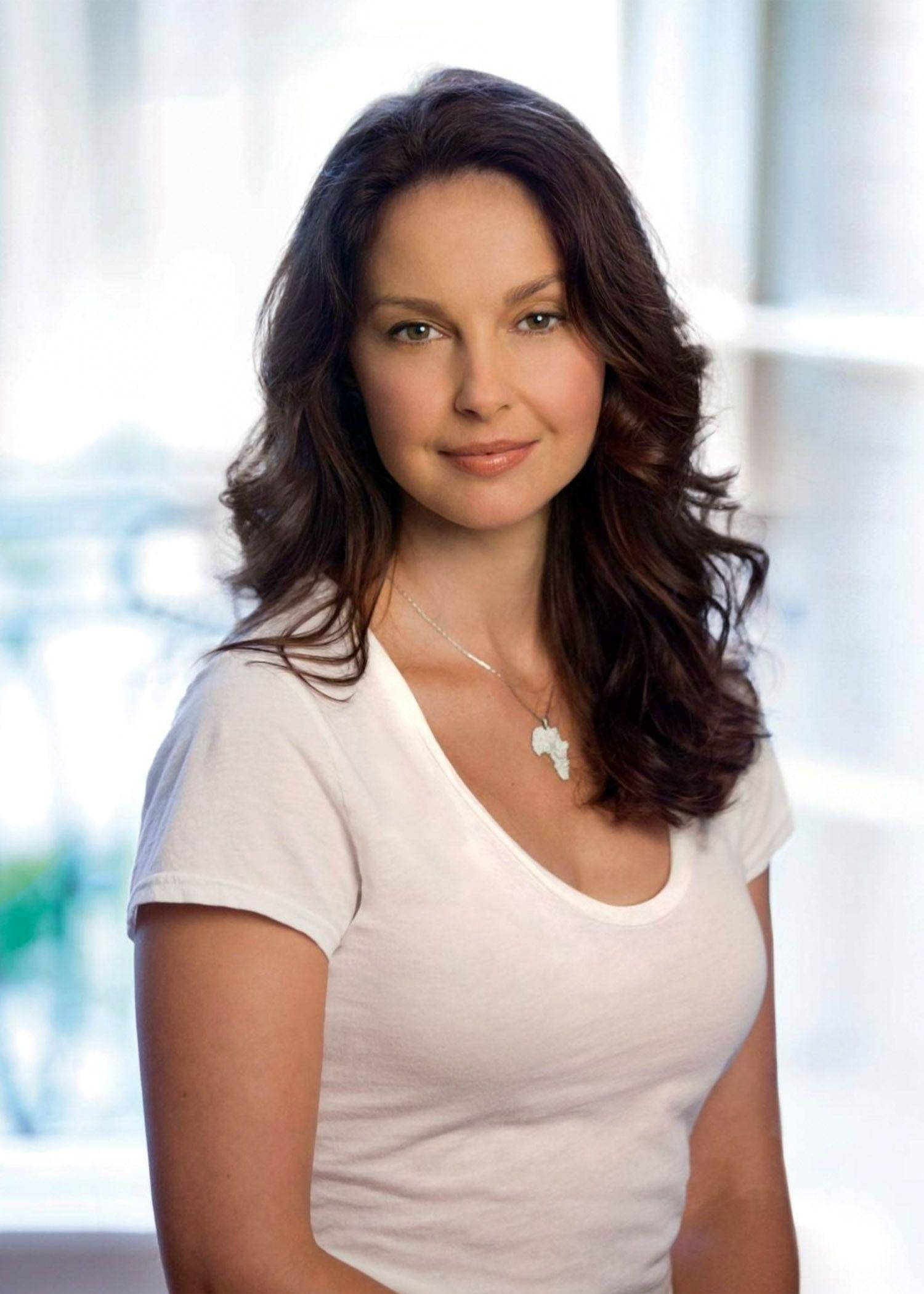 Ashley Judd 桌布 壁紙 — 一位擁有波浪棕色頭髮、身穿白色T恤的女性，在明亮模糊的窗戶背景下溫柔微笑。