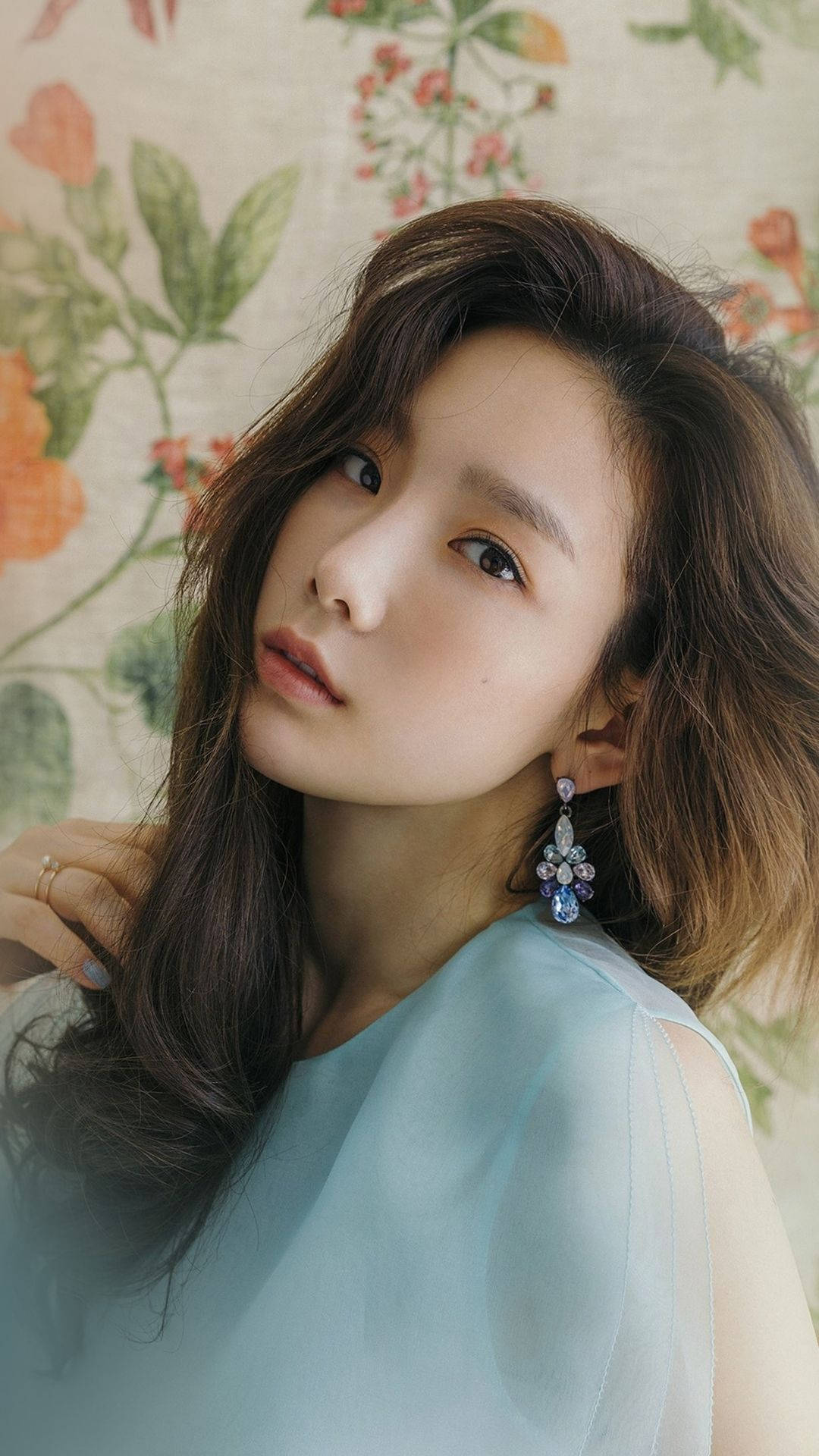 Taeyeon 桌布 壁紙 — 一位擁有波浪棕色頭髮和優雅藍色耳環的女性，在花卉壁紙背景下溫柔地凝視。