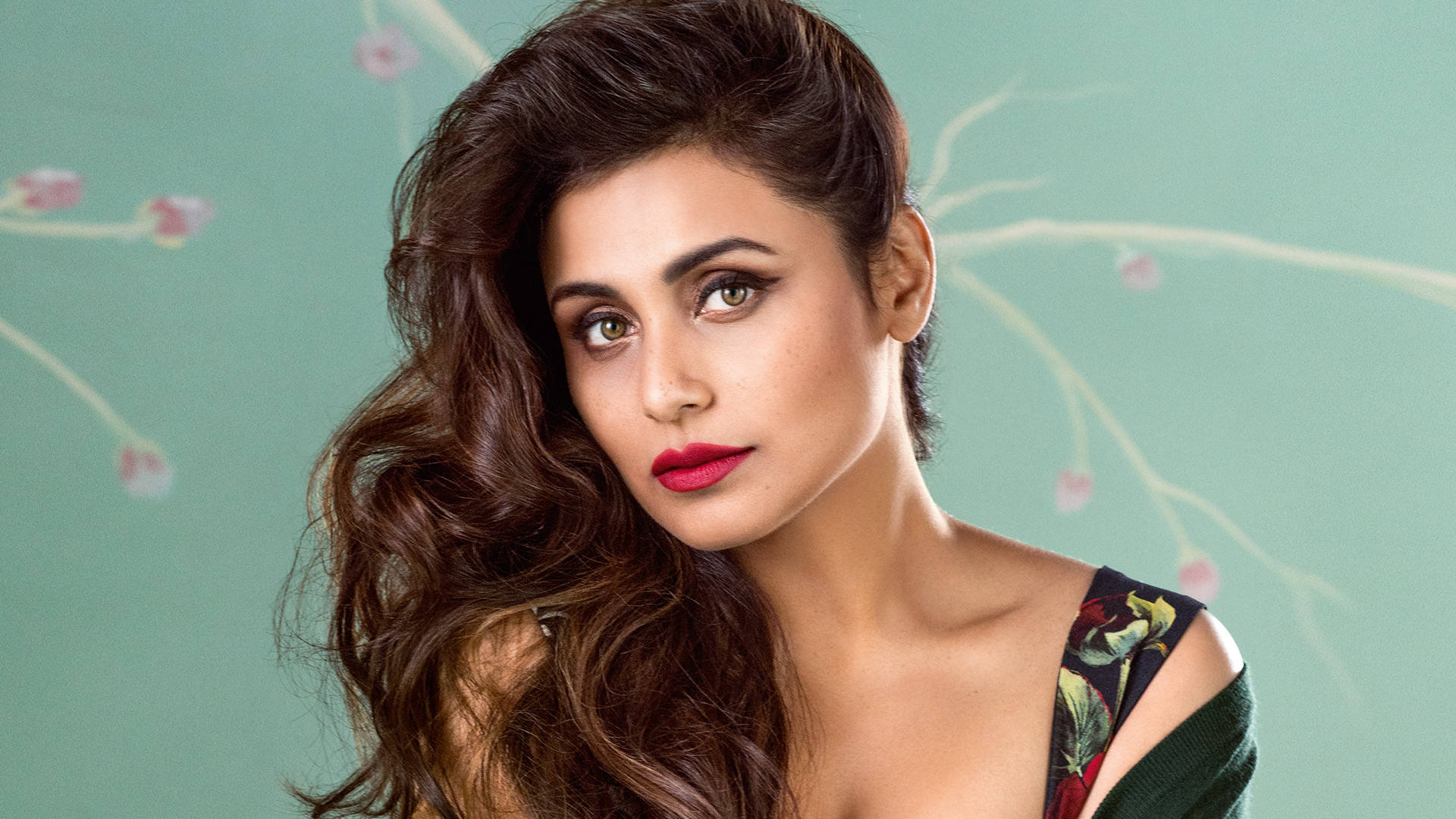 Rani Mukerji 桌布 壁紙 — 一位擁有波浪棕色頭髮和大膽紅唇的女性，直視著柔和的青綠色背景，背景點綴著精緻的花枝。
