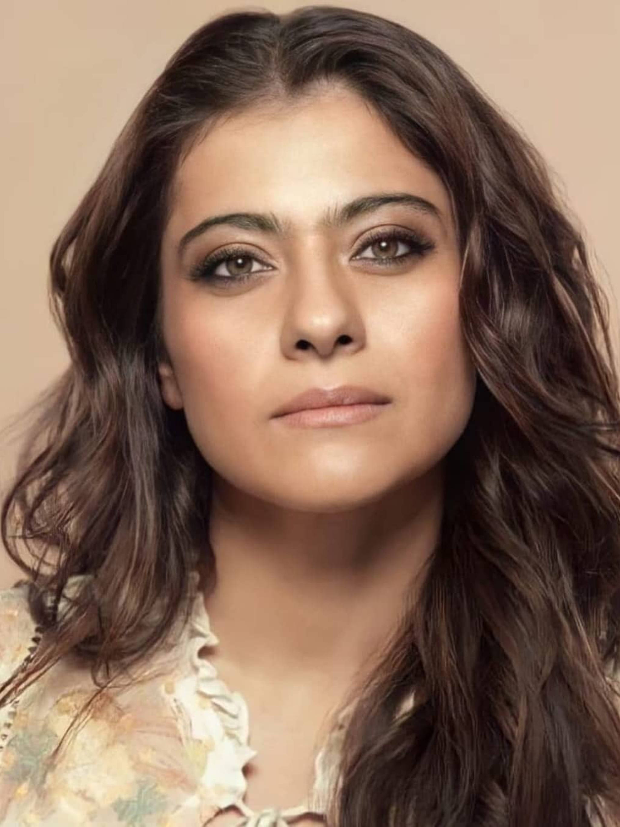 Kajol 桌布 壁紙 — 一位擁有波浪棕色頭髮和柔和妝容的女性在溫暖米色背景下的特寫肖像。