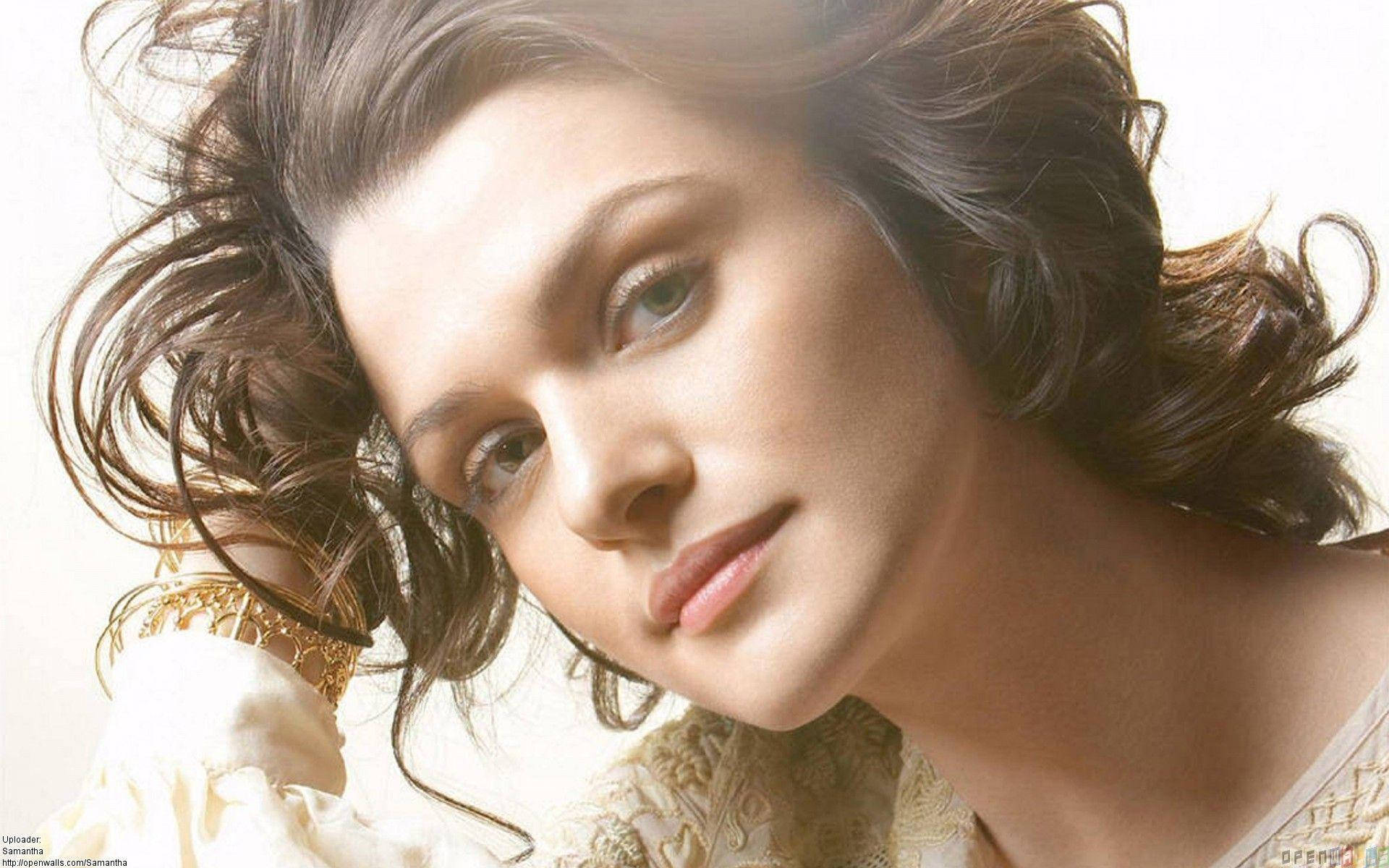 Rachel Weisz 桌布 壁紙 — 一位擁有波浪棕色頭髮和柔和目光的女性的特寫肖像，在明亮的白色背景前佩戴華麗的金色珠寶。