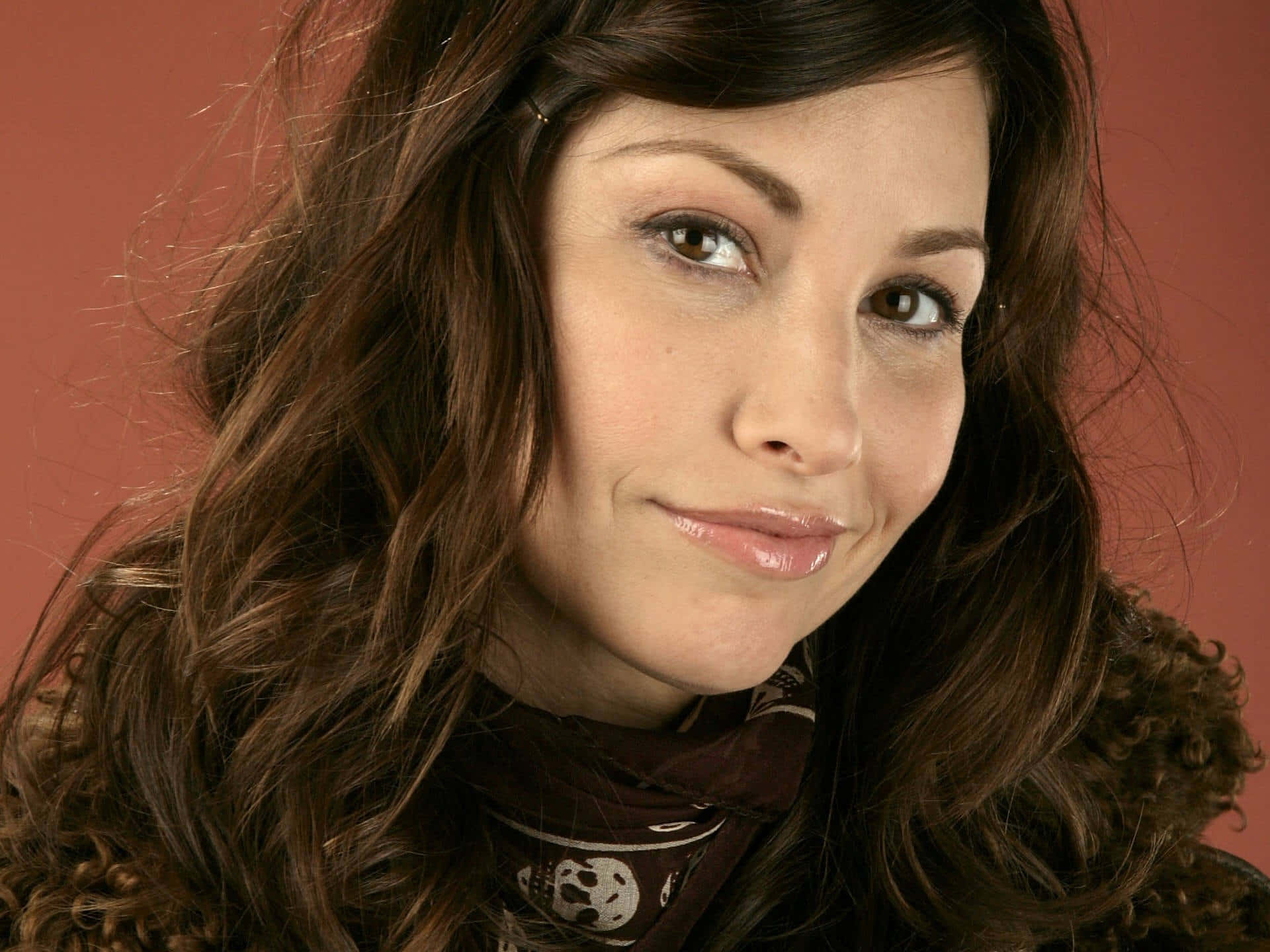 Gina Gershon 桌布 壁紙 — 一位擁有波浪棕色頭髮和溫柔微笑的女性的特寫肖像，背景為溫暖的陶土色。