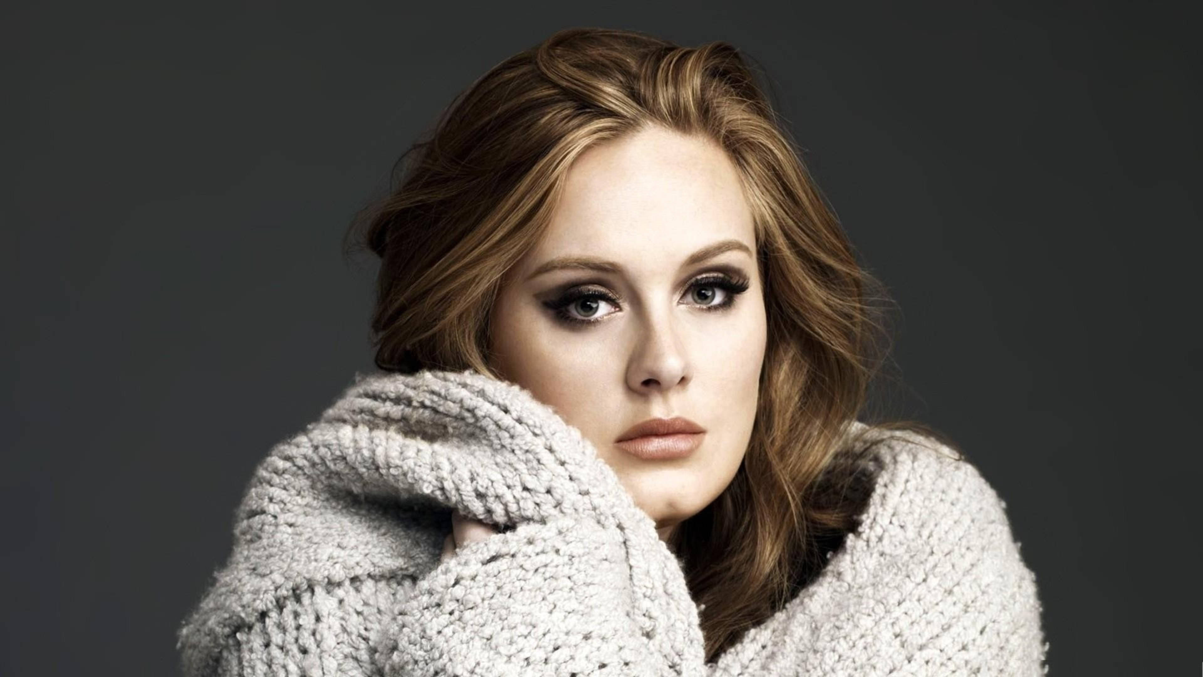Adele 桌布 壁紙 — 一位擁有波浪棕色頭髮和煙熏眼妝的女性，在深灰色背景下裹著厚實的針織毛衣，凝視著鏡頭。