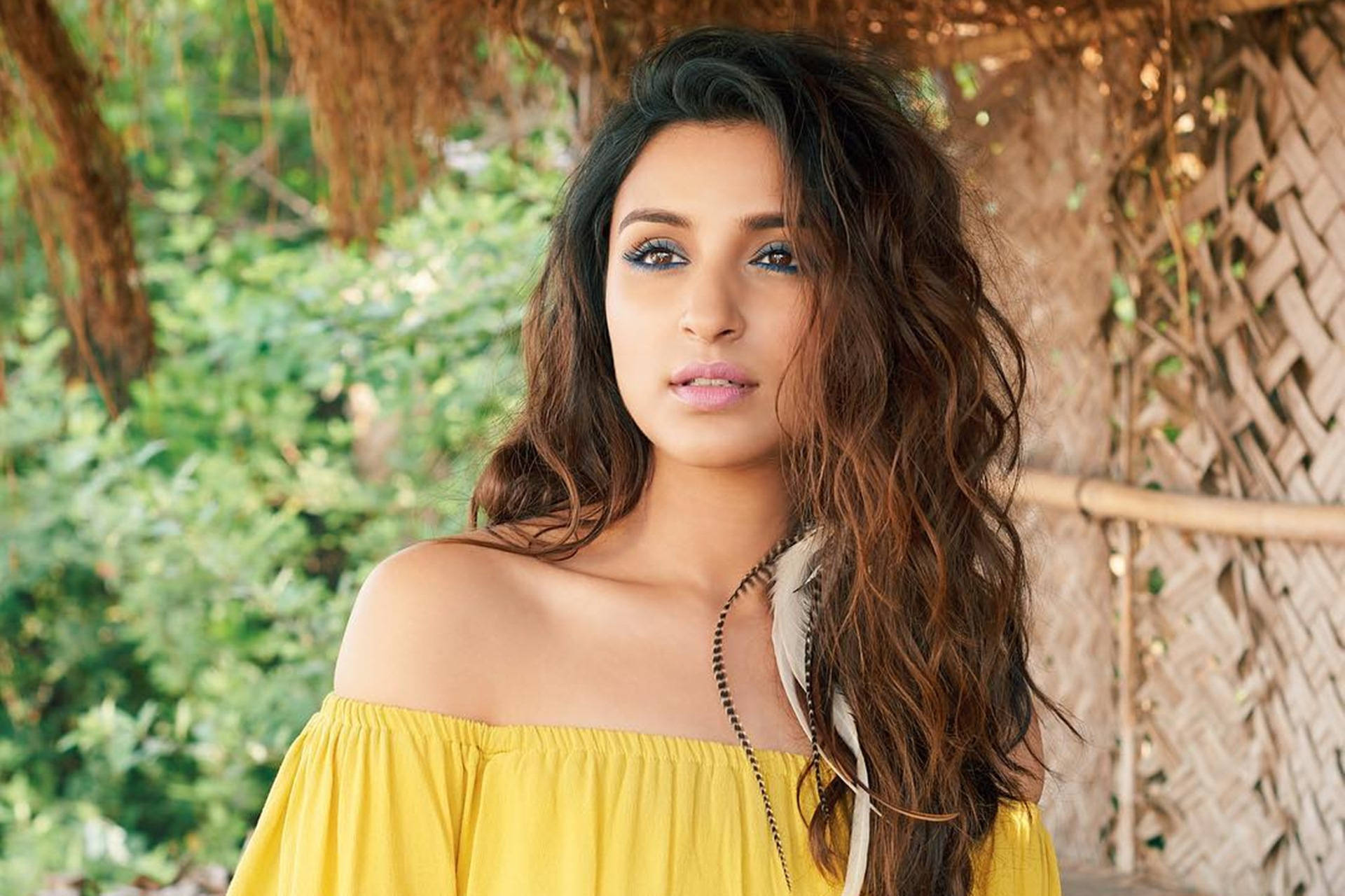 Parineeti 桌布 壁紙 — 一位擁有波浪棕色頭髮和粗眼線的女性，在茅草屋頂下穿著鮮黃色露肩上衣，周圍環繞著茂密的綠色植物。