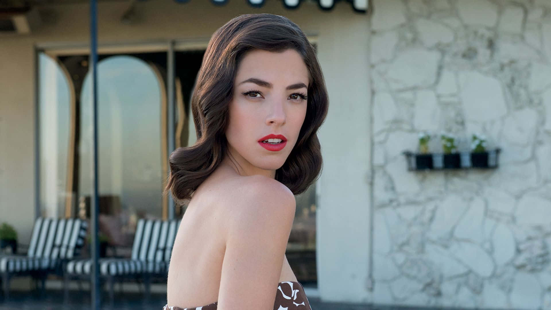 Olivia Thirlby 桌布 壁紙 — 一位擁有波浪棕色頭髮和紅唇的女性，在石牆和庭院椅子的襯托下轉頭回望。