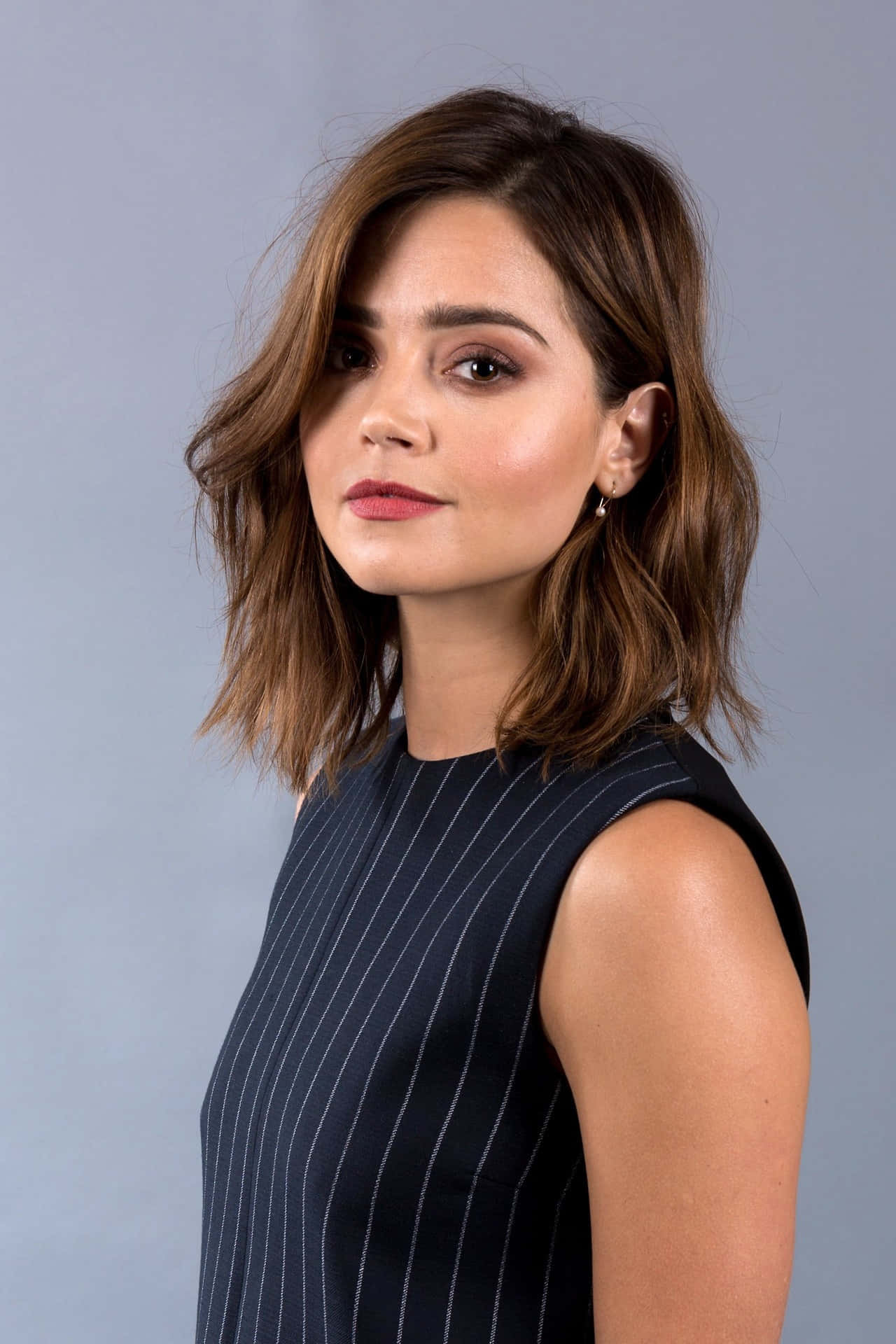 Jenna Coleman 桌布 壁紙 — 一位擁有波浪棕色頭髮和紅色唇膏的女性，在柔和灰色背景的攝影棚肖像中平靜地凝視。