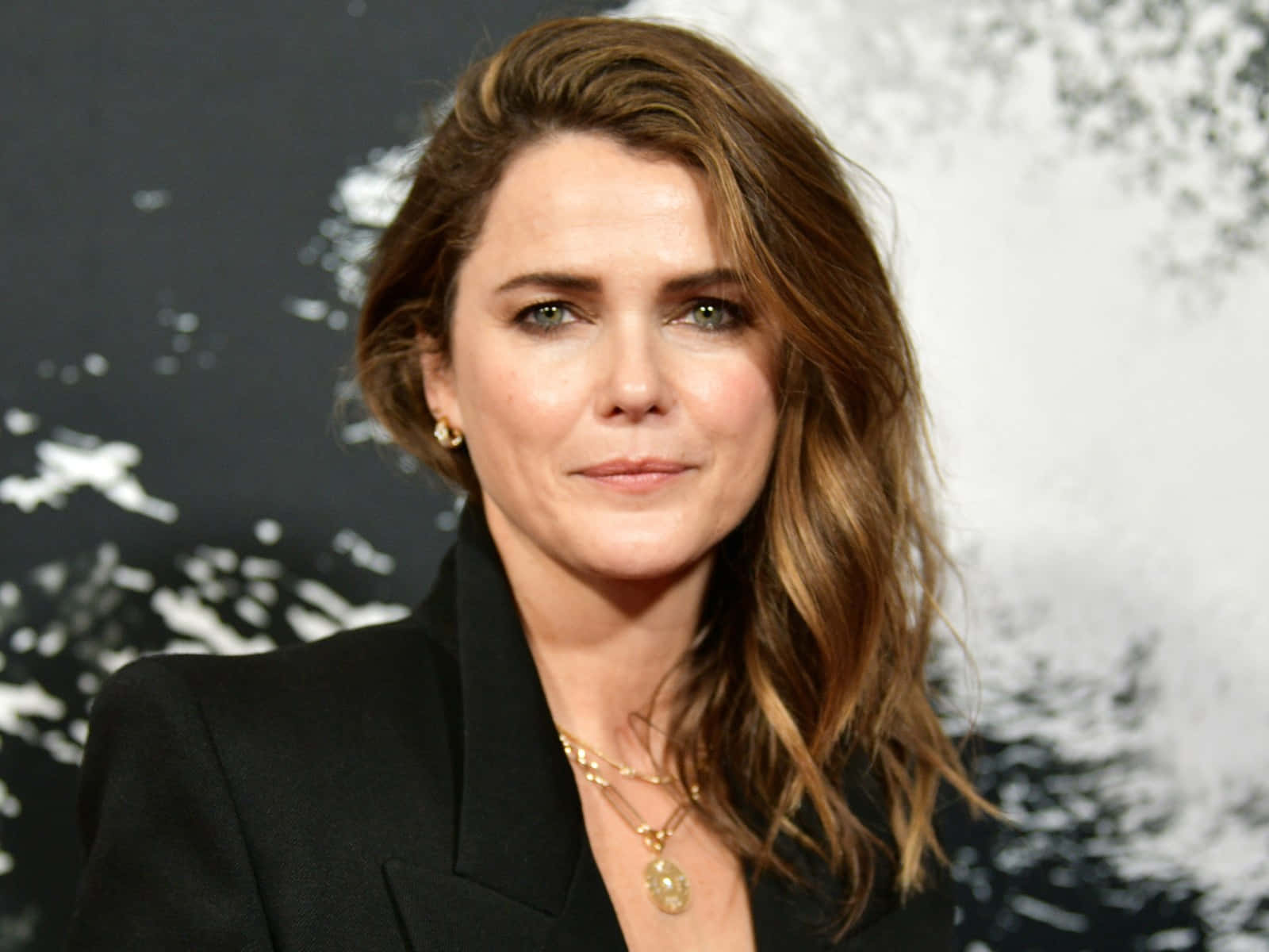 keri russell 桌布 壁紙 — 一位擁有波浪棕色頭髮和綠色眼睛的女性，在深色抽象背景前穿著黑色西裝外套和金色珠寶。