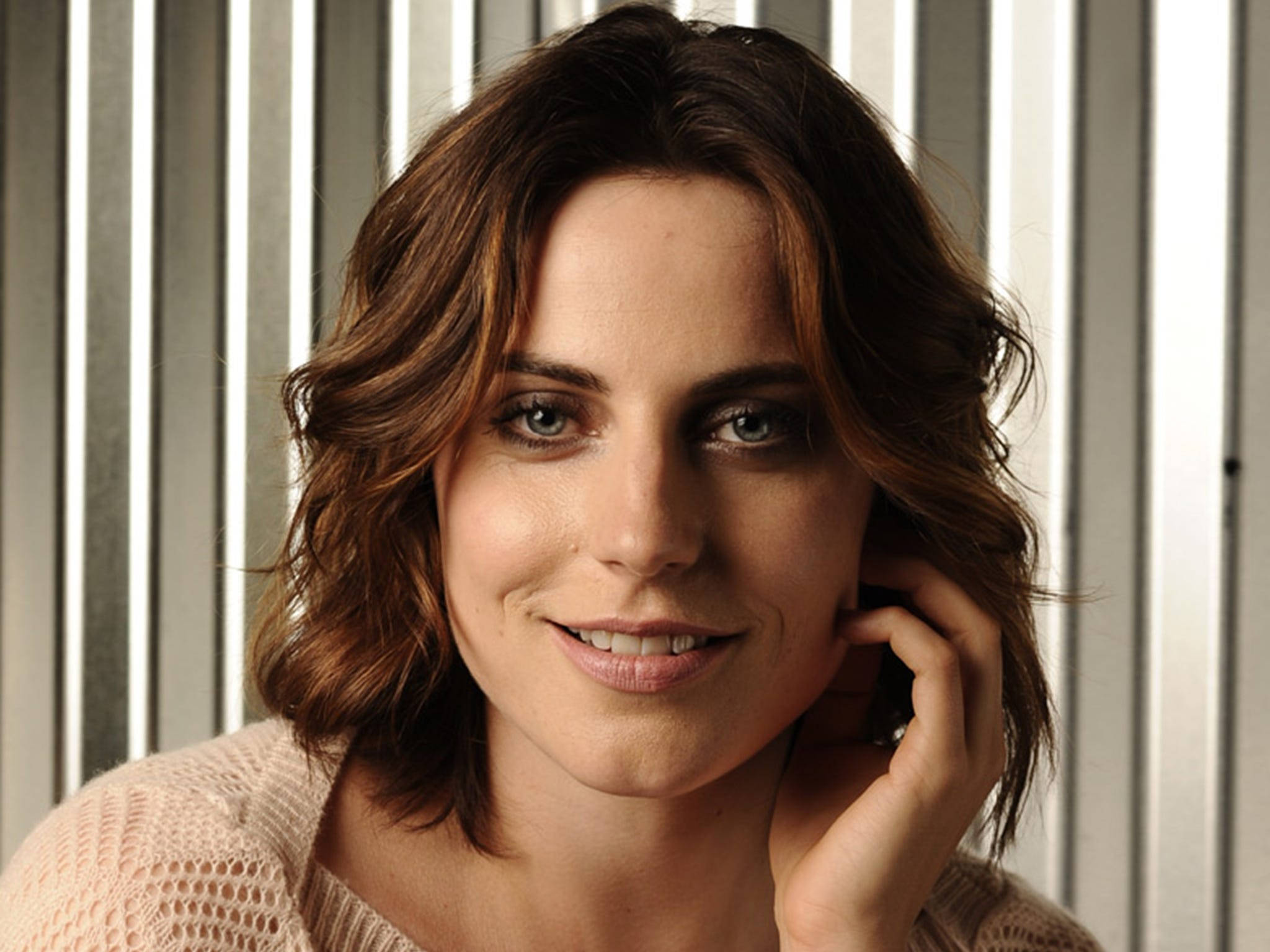 Antje Traue 桌布 壁紙 — 一位擁有波浪棕色頭髮和藍色眼睛的女性，將臉頰貼在條紋背景上，溫柔地微笑。