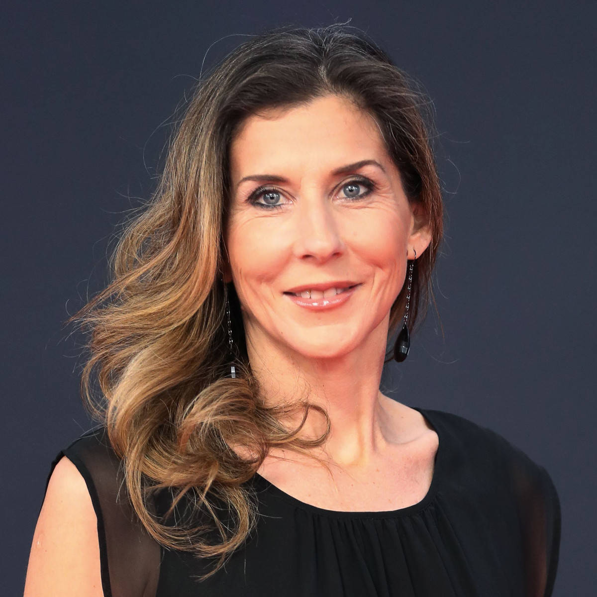 Monica Seles 桌布 壁紙 — 一位擁有波浪棕色頭髮和藍色眼睛的女性，在深藍色背景下微笑，穿著黑色上衣和垂墜耳環。