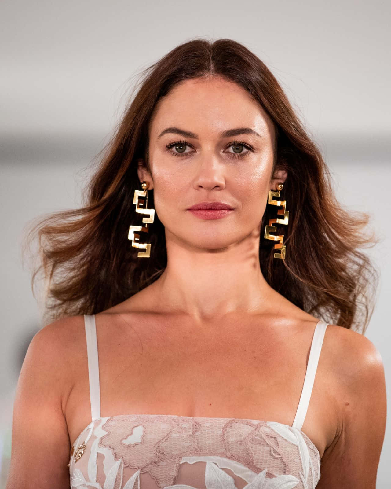Olga Kurylenko 桌布 壁紙 — 一位擁有波浪棕色頭髮和醒目幾何金色耳環的女性，穿著白色蕾絲上衣直視鏡頭。