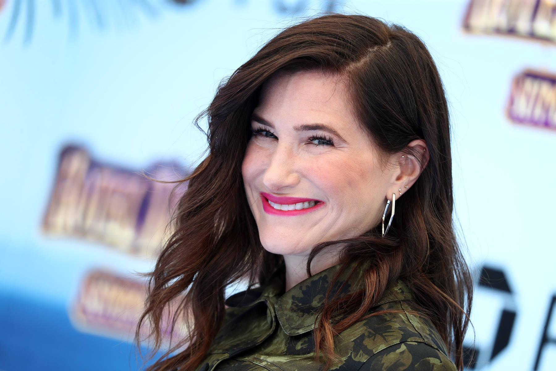 Celebrity 桌布 壁紙 — 一位擁有波浪棕色頭髮和鮮紅色唇膏的女性，穿著迷彩外套，溫暖地微笑。
