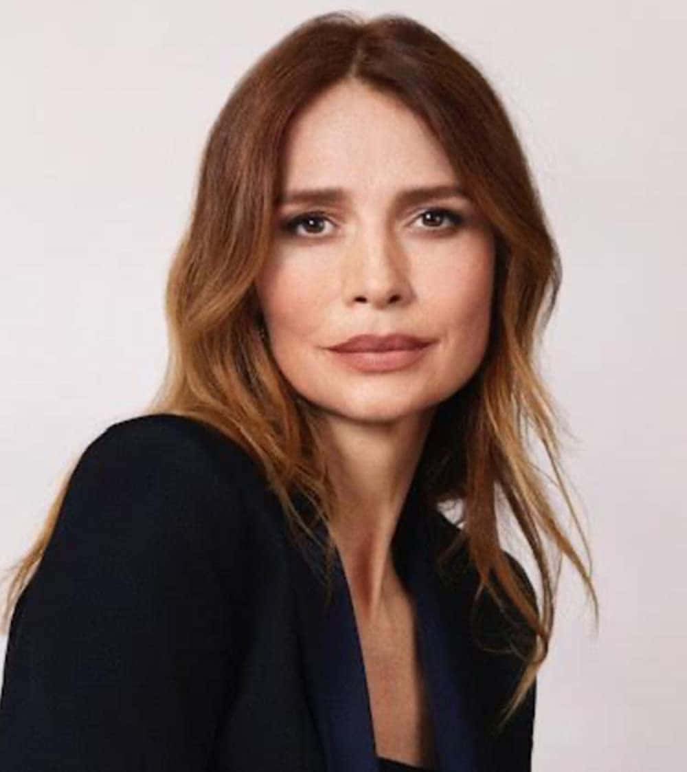 Saffron Burrows 桌布 壁紙 — 一位擁有波浪棕色頭髮和黑色西裝外套的女性，在純白色背景前平靜地凝視著相機。