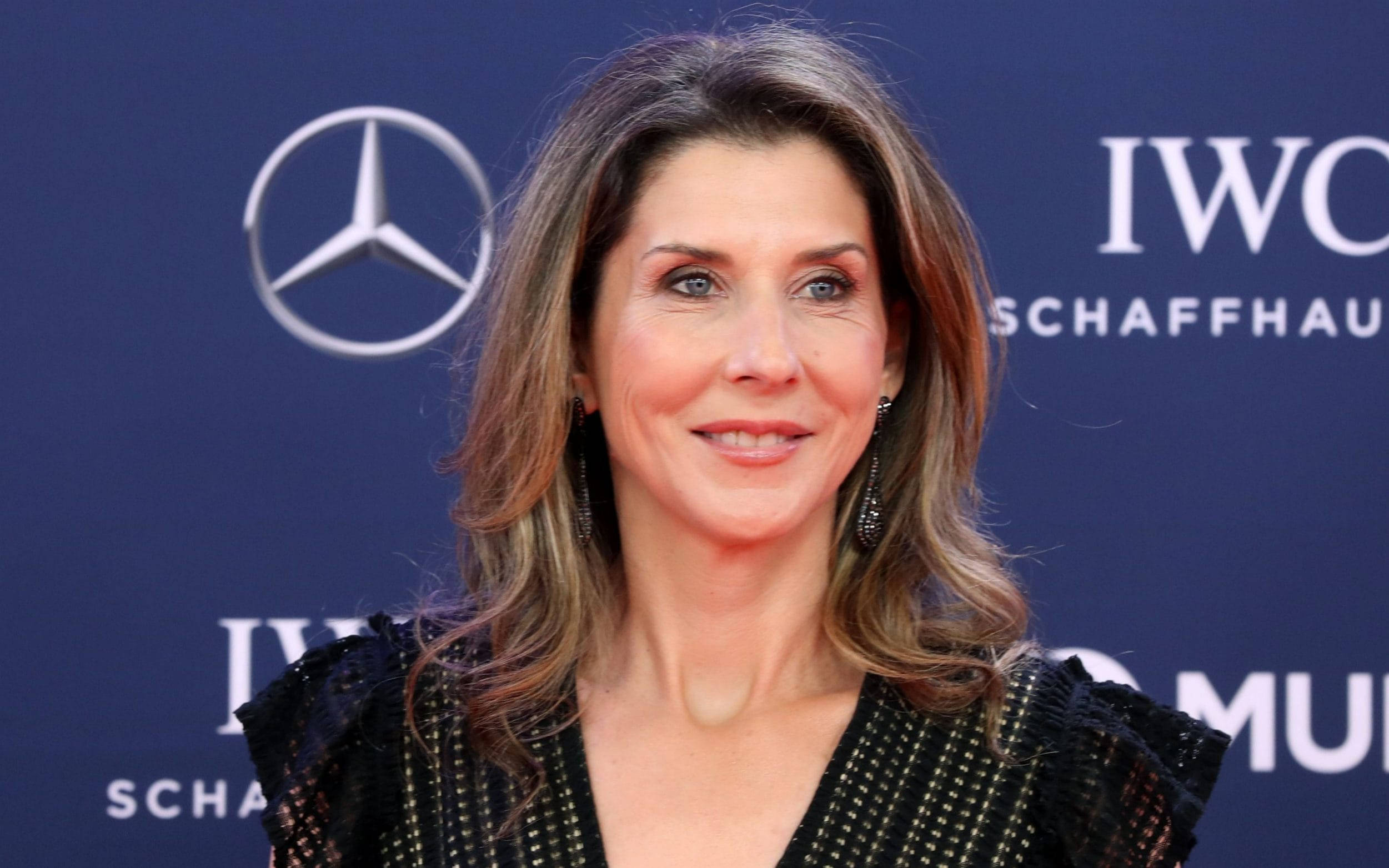 Monica Seles 桌布 壁紙 — 一位擁有波浪棕色頭髮的女性，穿著黑色珠飾連衣裙，在藍色品牌背景前對著鏡頭微笑。