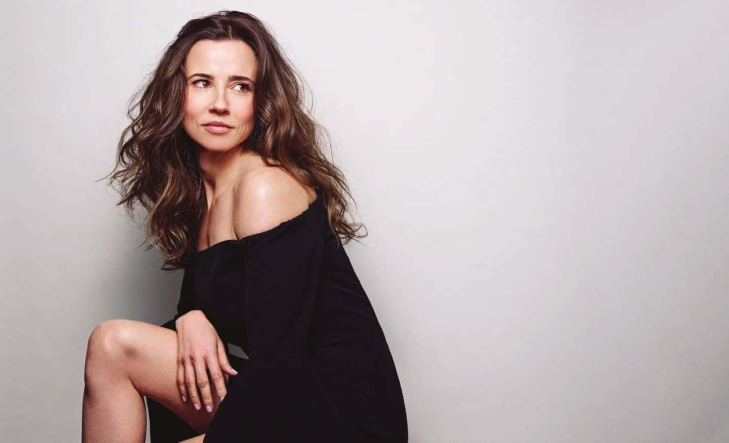 linda cardellini 桌布 壁紙 — 一位擁有波浪棕色頭髮的女性，穿著黑色露肩連衣裙，坐在純白色背景前。