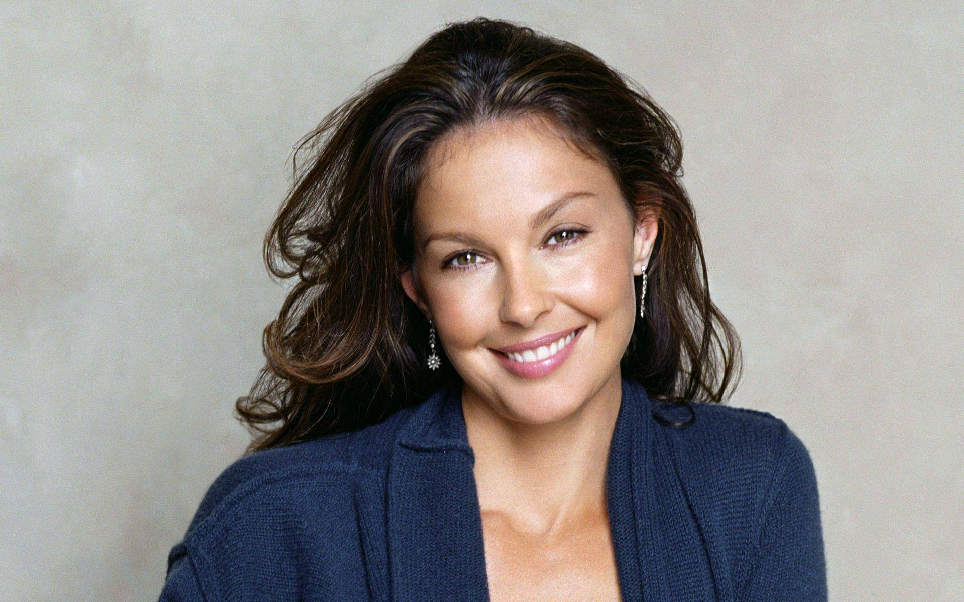 Ashley Judd 桌布 壁紙 — 一位擁有波浪棕色頭髮的女性，穿著海軍藍開襟毛衣，在中性紋理背景前溫暖地微笑。