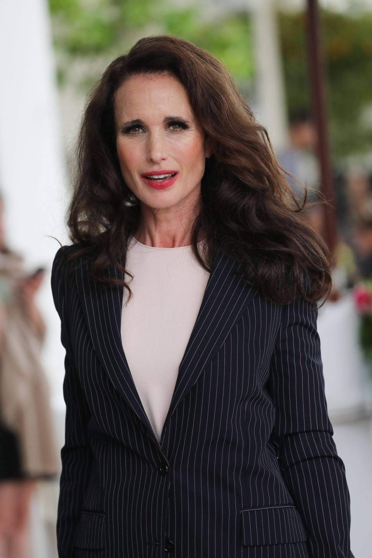 andie macdowell 桌布 壁紙 — 一位擁有波浪棕色頭髮的女性，身穿深色細條紋西裝外套，內搭淺色上衣，在戶外面對鏡頭微笑。