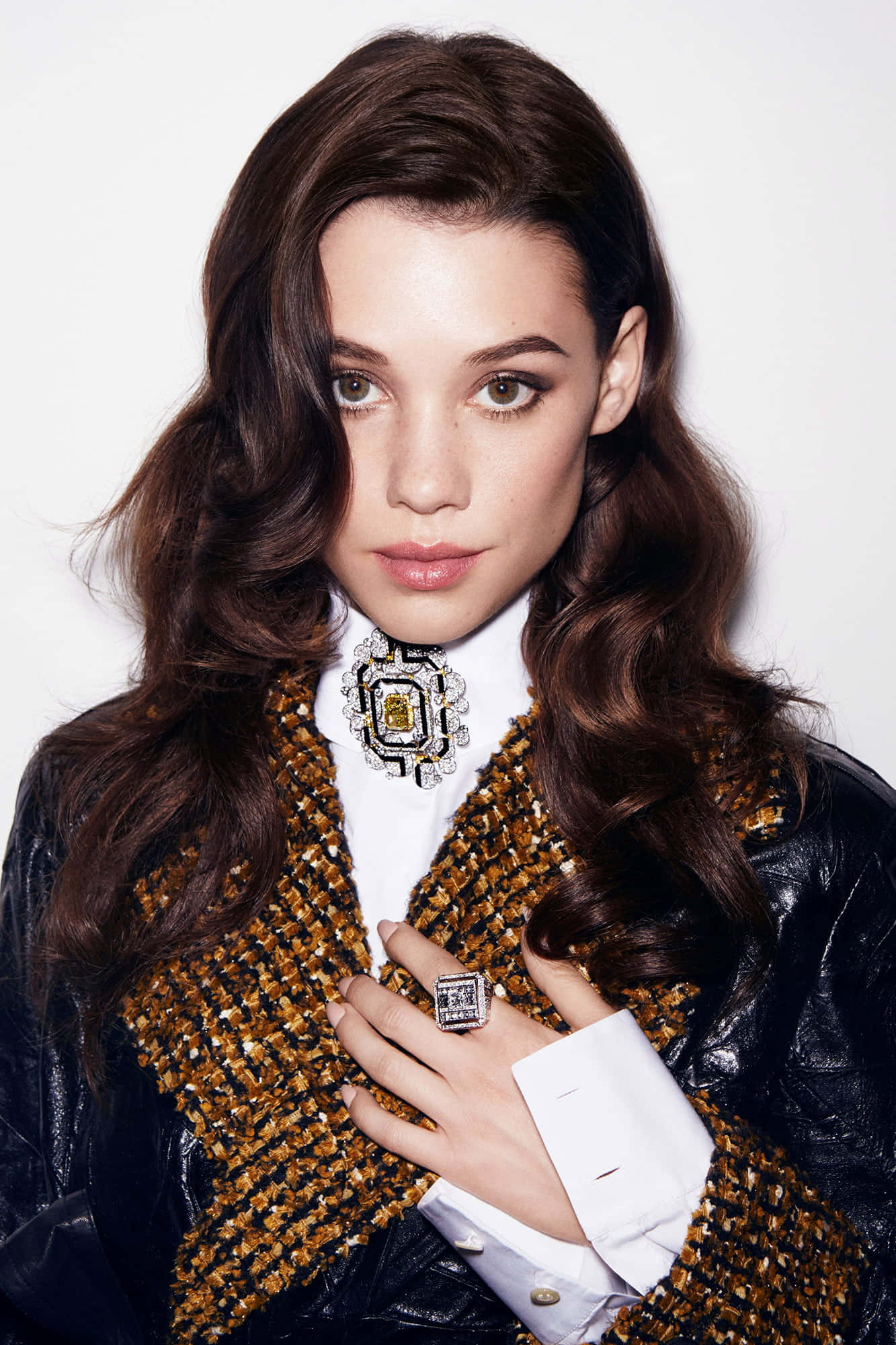 Astrid Berges Frisbey 桌布 壁紙 — 一位擁有波浪棕色頭髮的女性，身穿黑色皮夾克，內搭金色粗花呢背心和白色襯衫，佩戴大型幾何胸針和相配的戒指。