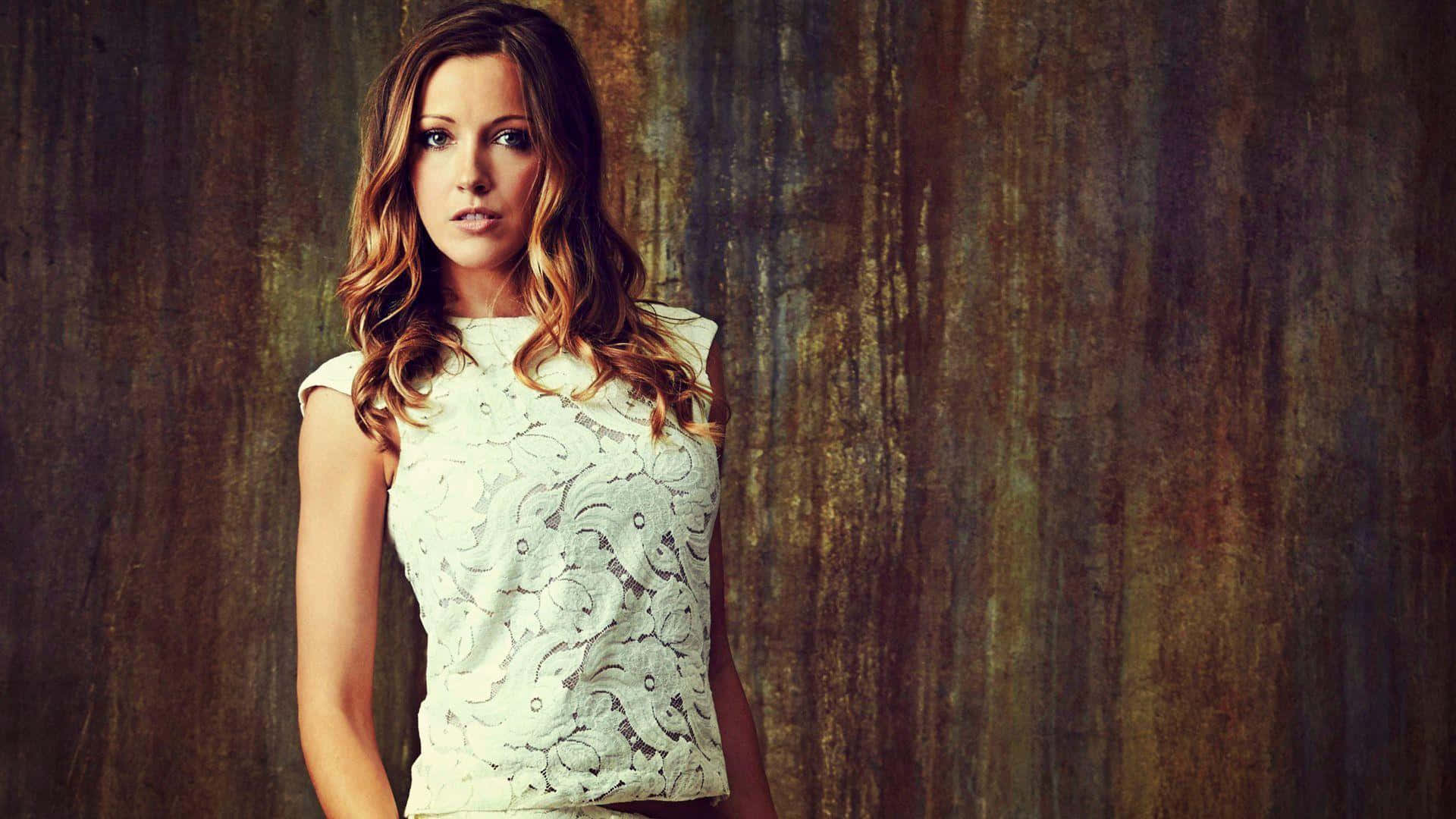 katie cassidy 桌布 壁紙 — 一位擁有波浪棕色頭髮的女性，身穿白色蕾絲上衣，背景是帶有紋理的鏽褐色牆壁。