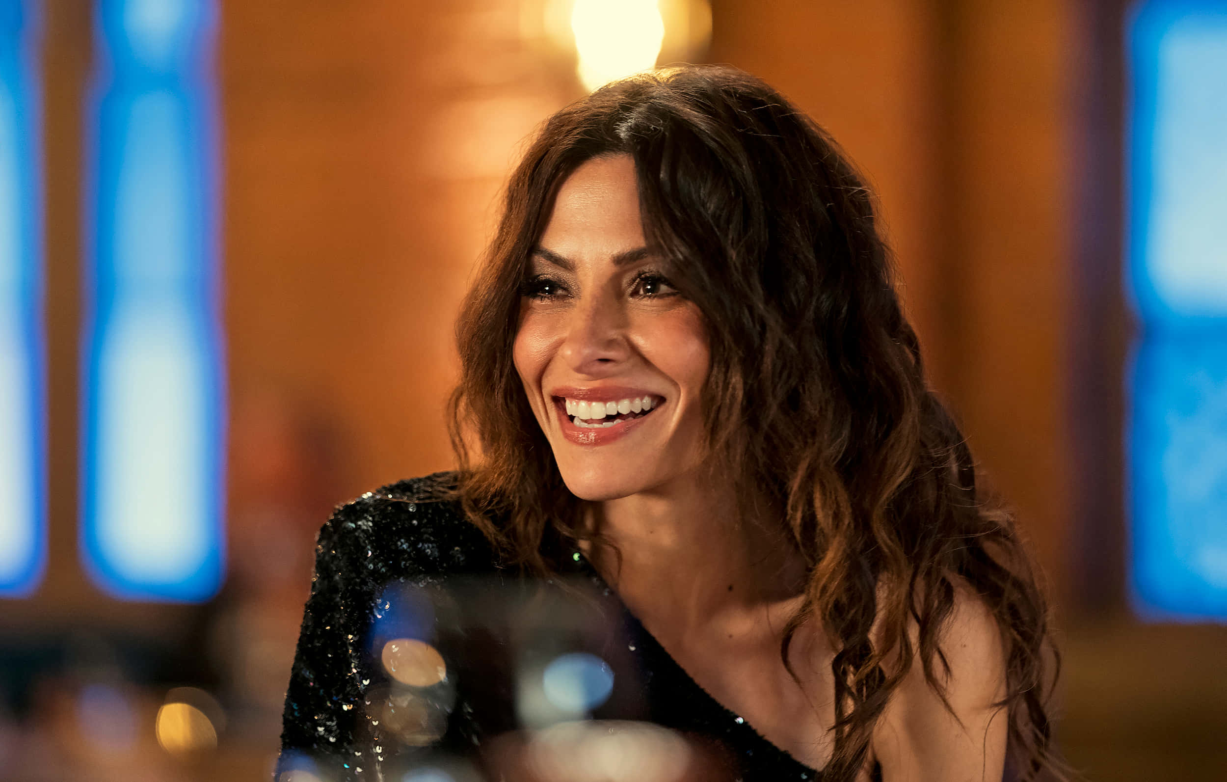 sarah shahi 桌布 壁紙 — 一位擁有波浪棕色頭髮的女性在昏暗的房間裡，在藍色和橙色燈光下溫暖地微笑。