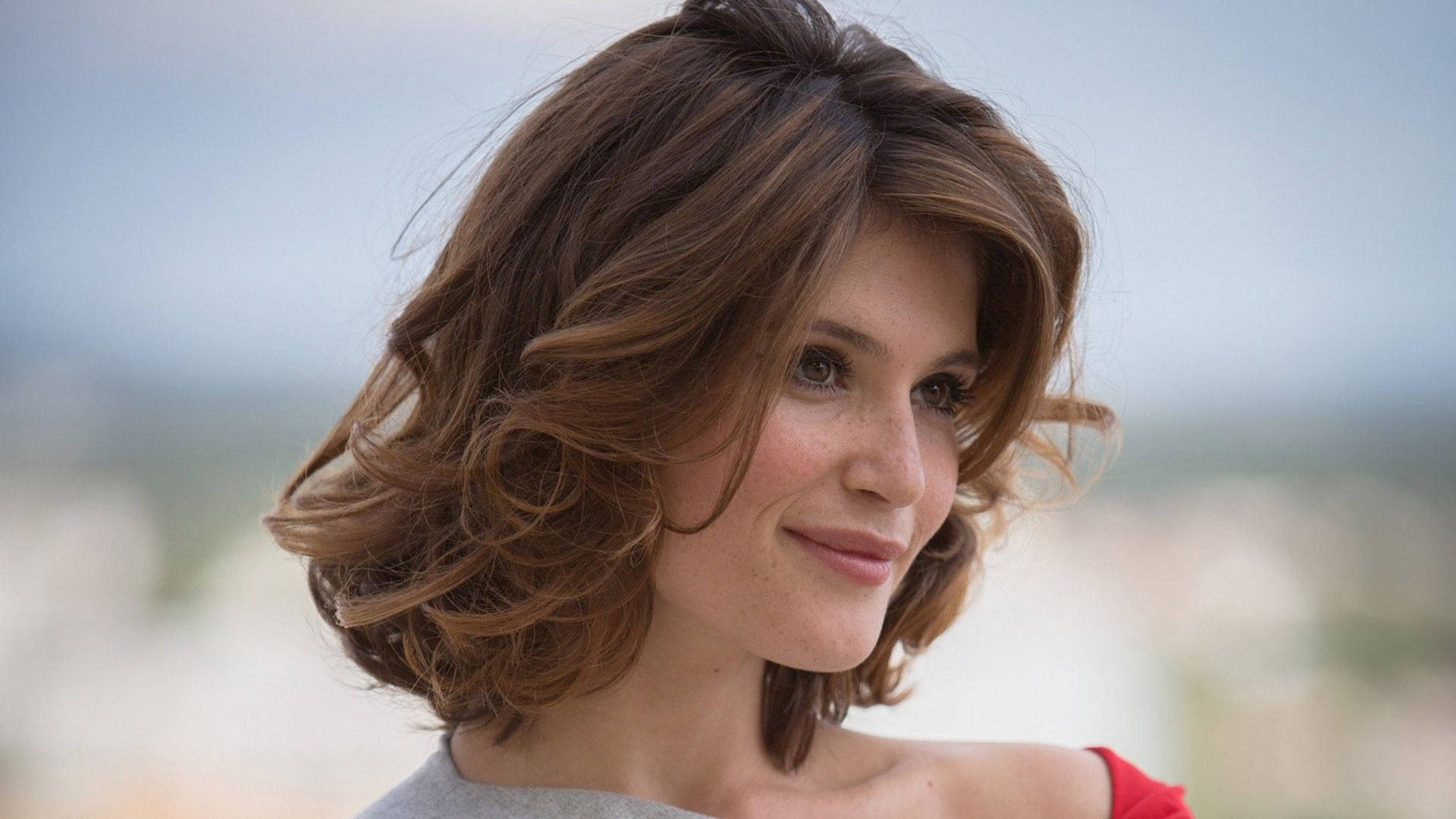 Gemma Arterton 桌布 壁紙 — 一位擁有波浪棕色頭髮的女性在模糊的城市景觀背景下溫柔微笑。