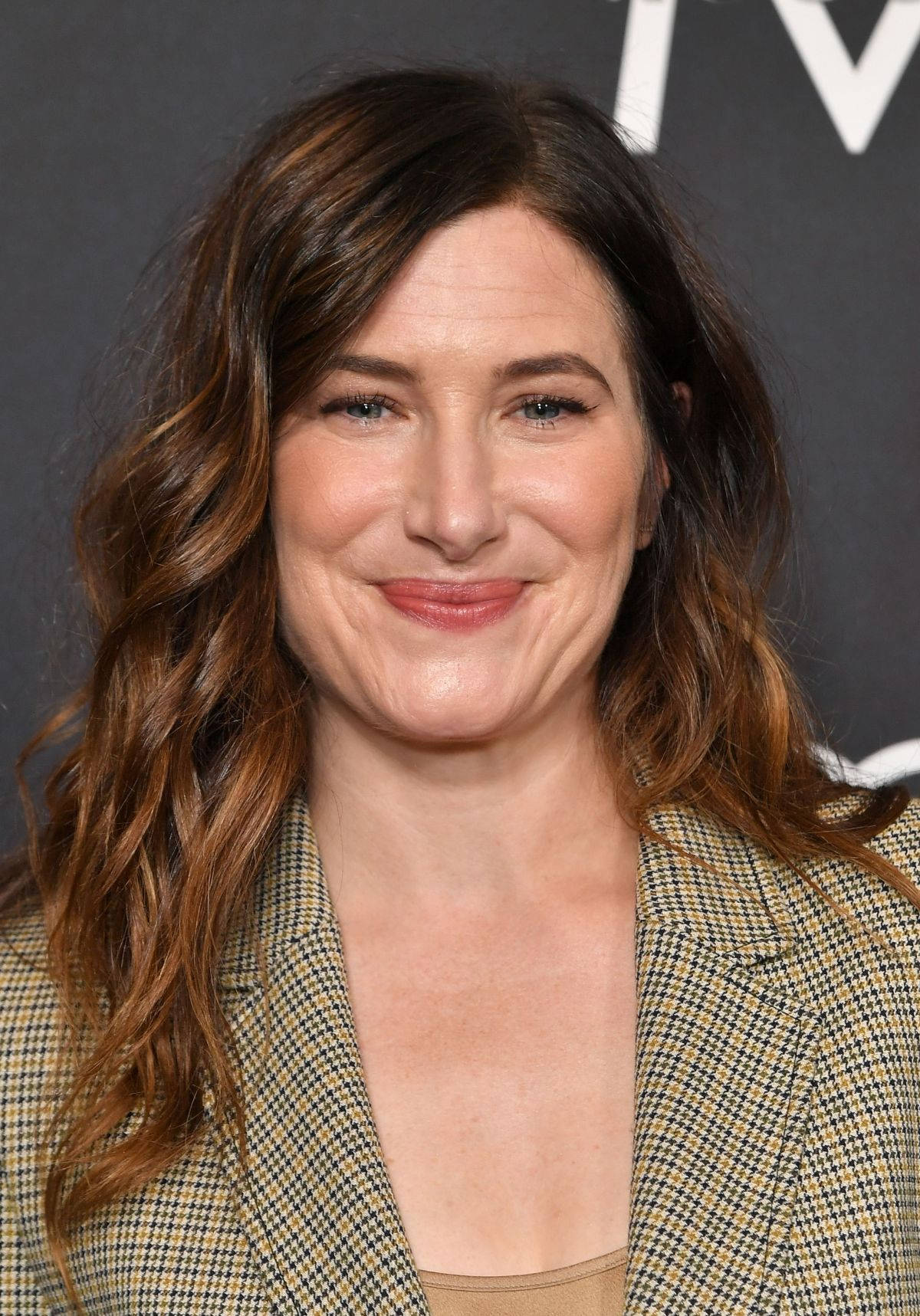 Celebrity 桌布 壁紙 — 一位擁有波浪棕色頭髮的女性在深色背景下穿著格紋西裝外套溫暖微笑的特寫肖像。