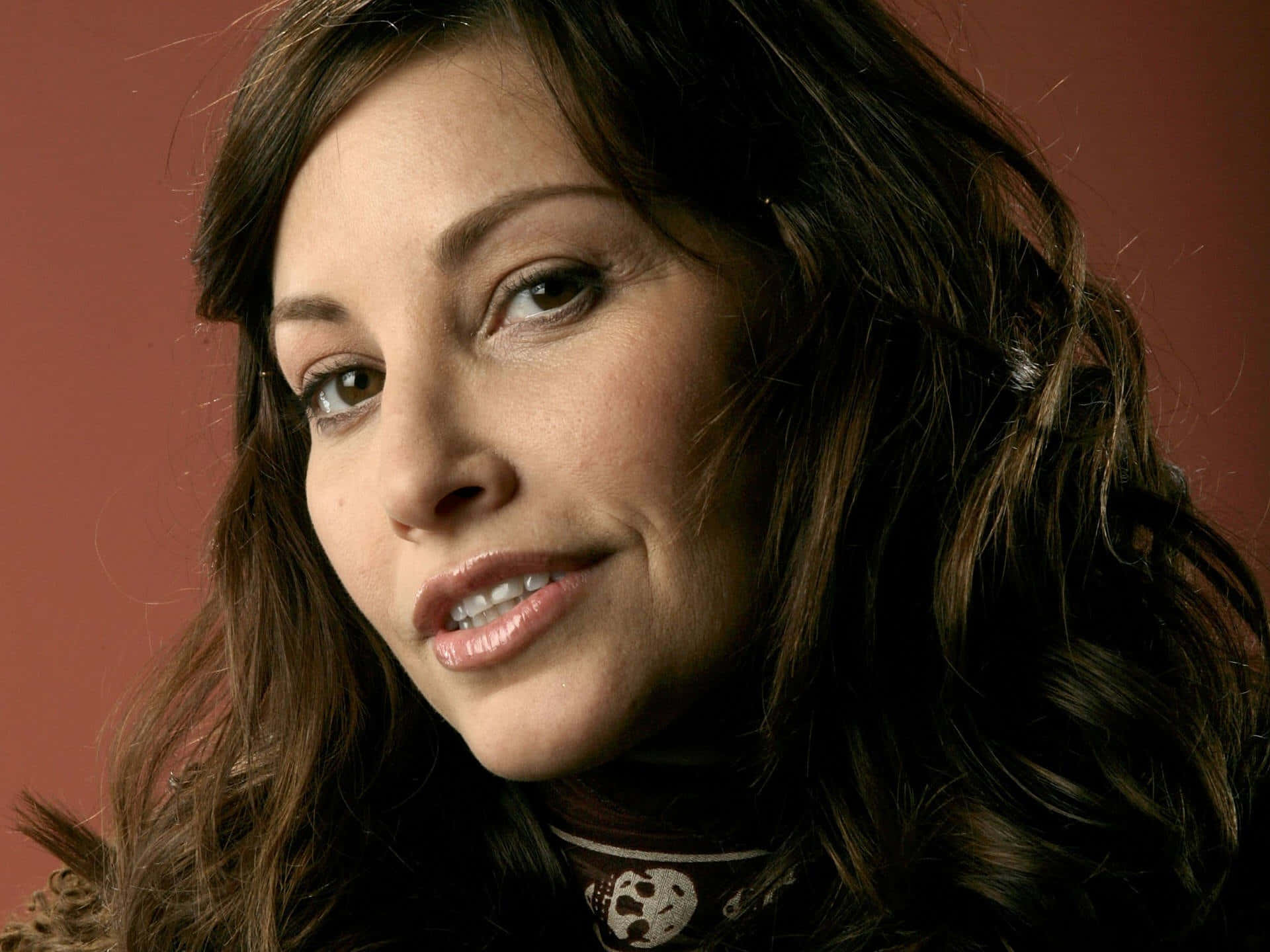 Gina Gershon 桌布 壁紙 — 一位擁有波浪棕色頭髮的女性在溫暖的赤陶色背景前微笑的特寫肖像。
