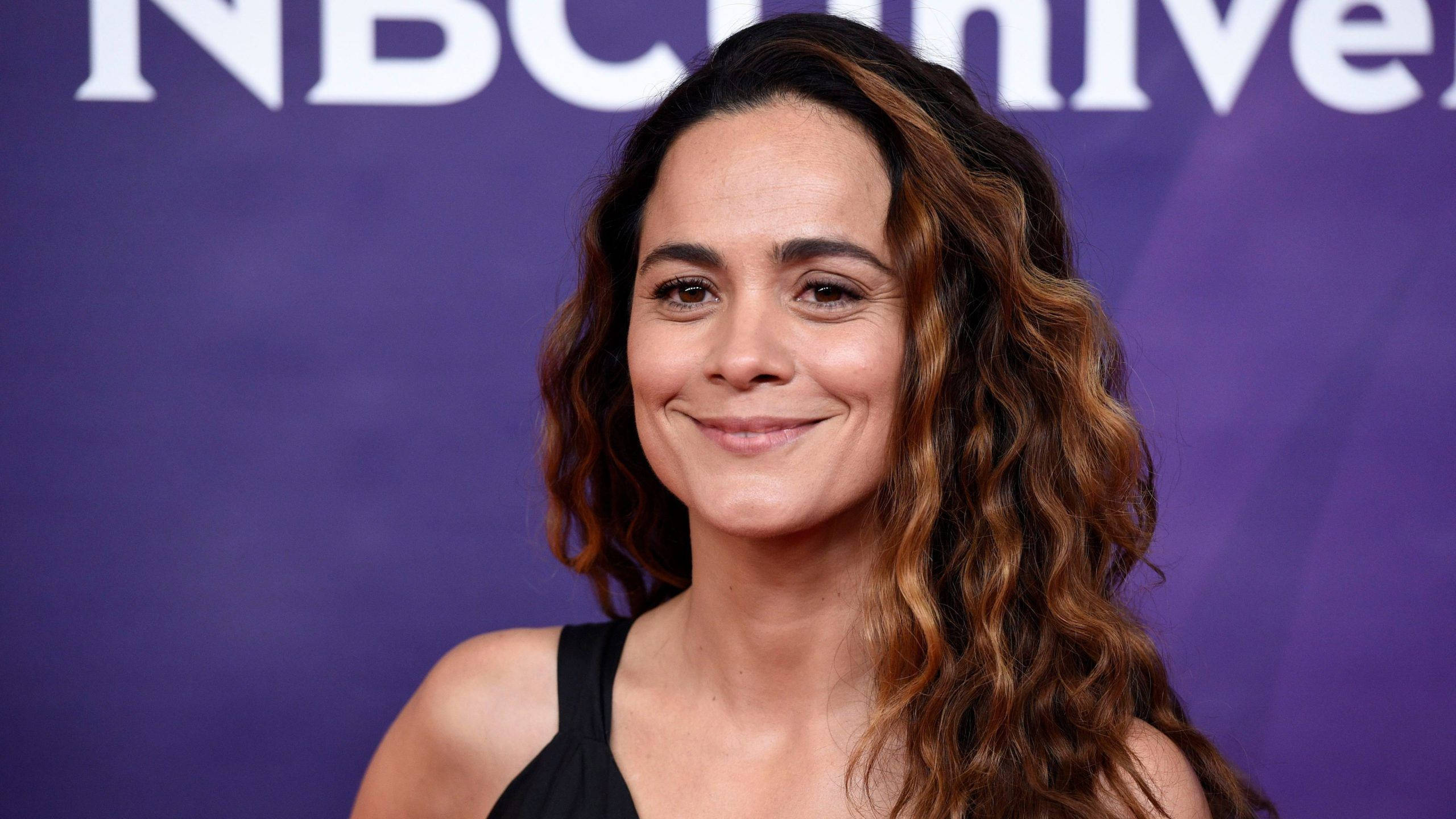 Celebrity 桌布 壁紙 — 一位擁有波浪棕色頭髮的女性在紫色NBCUniversal背景前溫暖微笑。