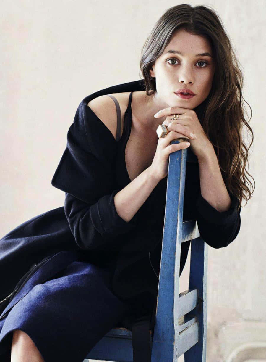Astrid Berges Frisbey 桌布 壁紙 — 一位擁有波浪棕色頭髮的女性坐在藍色木椅上，穿著黑色露肩連衣裙，直視鏡頭。