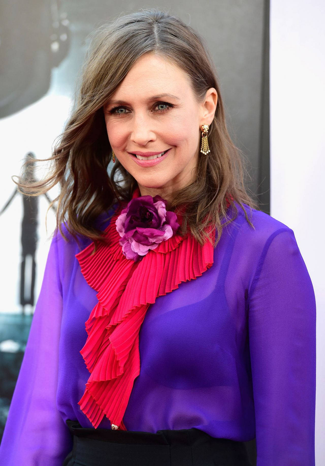 Celebrity 桌布 壁紙 — 一位擁有波浪棕色頭髮的女性溫暖微笑，身穿鮮豔紫色襯衫，搭配大膽的粉紅色荷葉邊領口和紫色花朵裝飾。