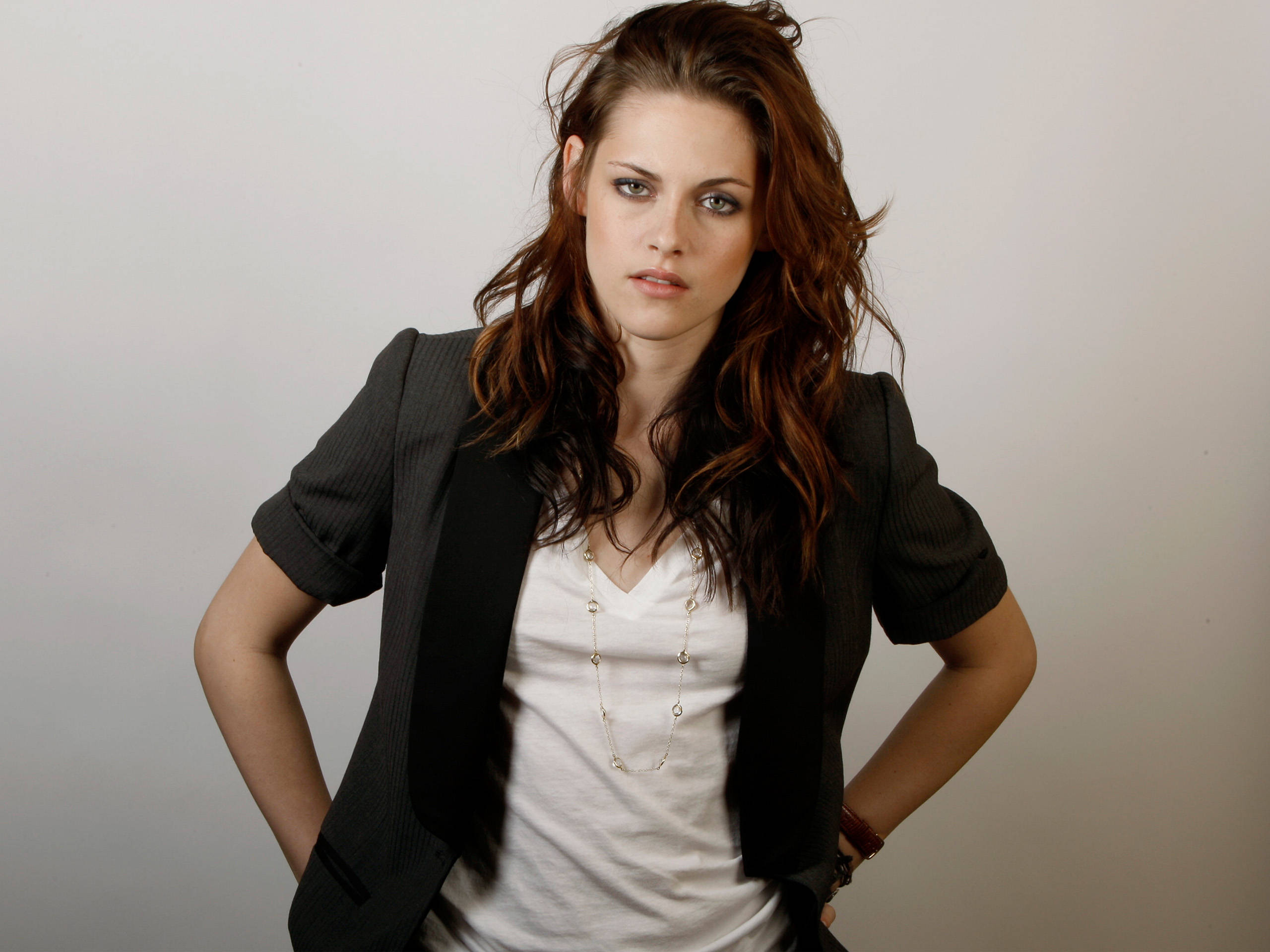 Kristen 桌布 壁紙 — 一位擁有波浪棕色頭髮的女性自信地站在純灰色背景前，身穿黑色西裝外套內搭白色T恤。