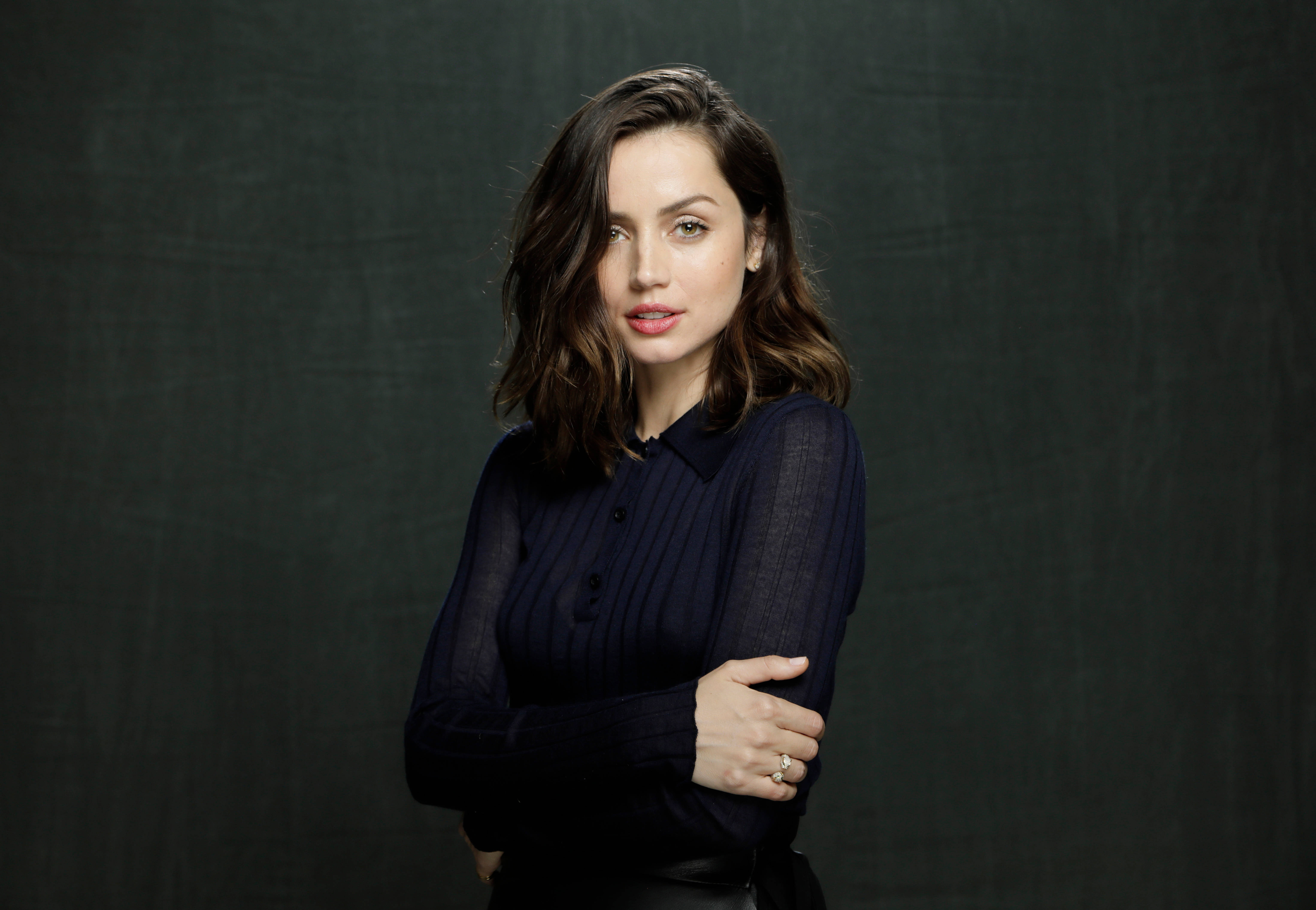 ana de armas 桌布 壁紙 — 一位擁有波浪棕色頭髮的女性自信地站在深色紋理背景前，穿著海軍藍羅紋上衣。