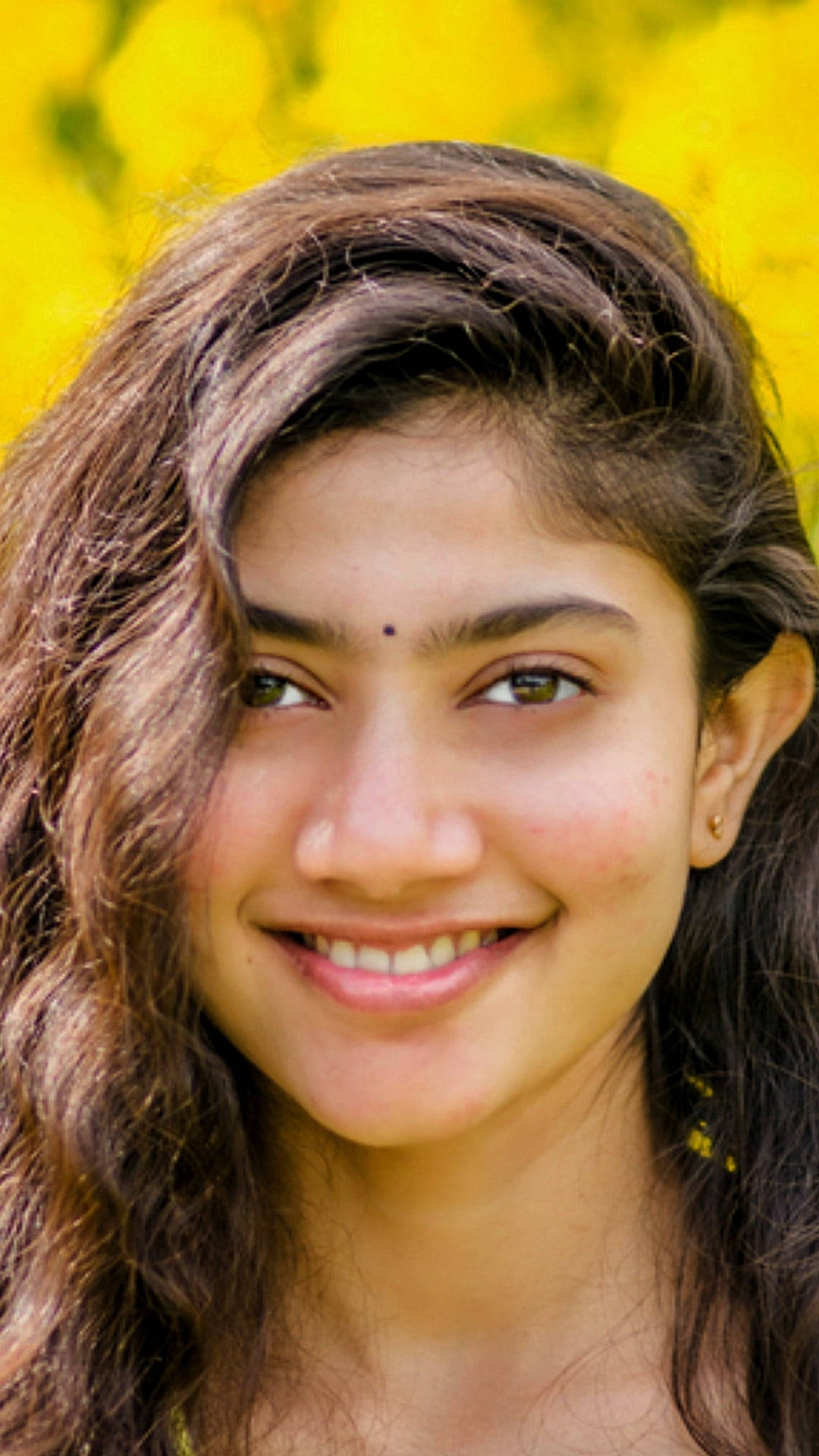 Sai Pallavi 桌布 壁紙 — 一位擁有波浪棕色頭髮的年輕女性在充滿活力的黃色花卉背景下溫暖地微笑。