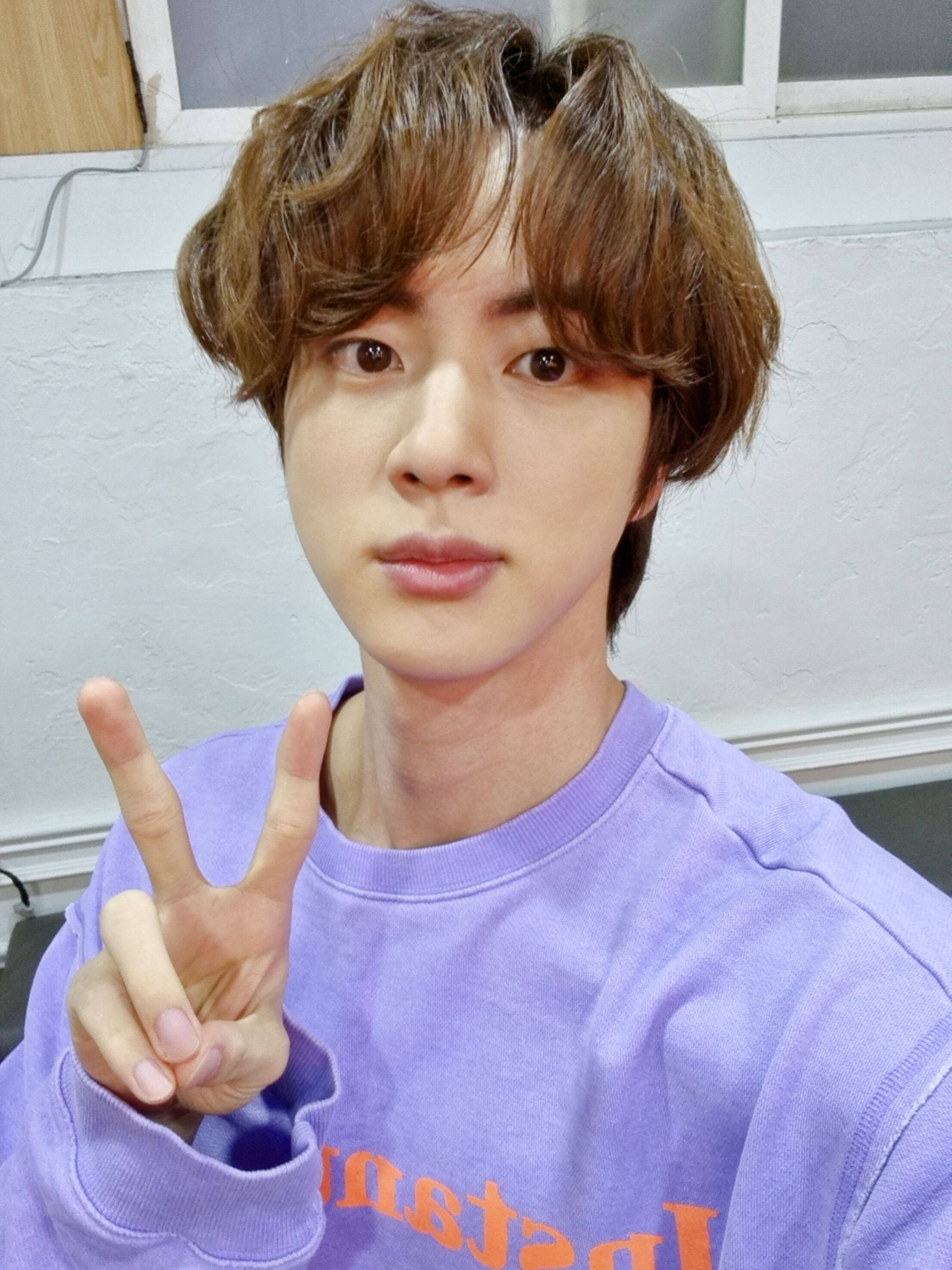 kim seok jin 桌布 壁紙 — 一位擁有波浪棕色頭髮的年輕男子穿著紫色運動衫，用手比出和平手勢。