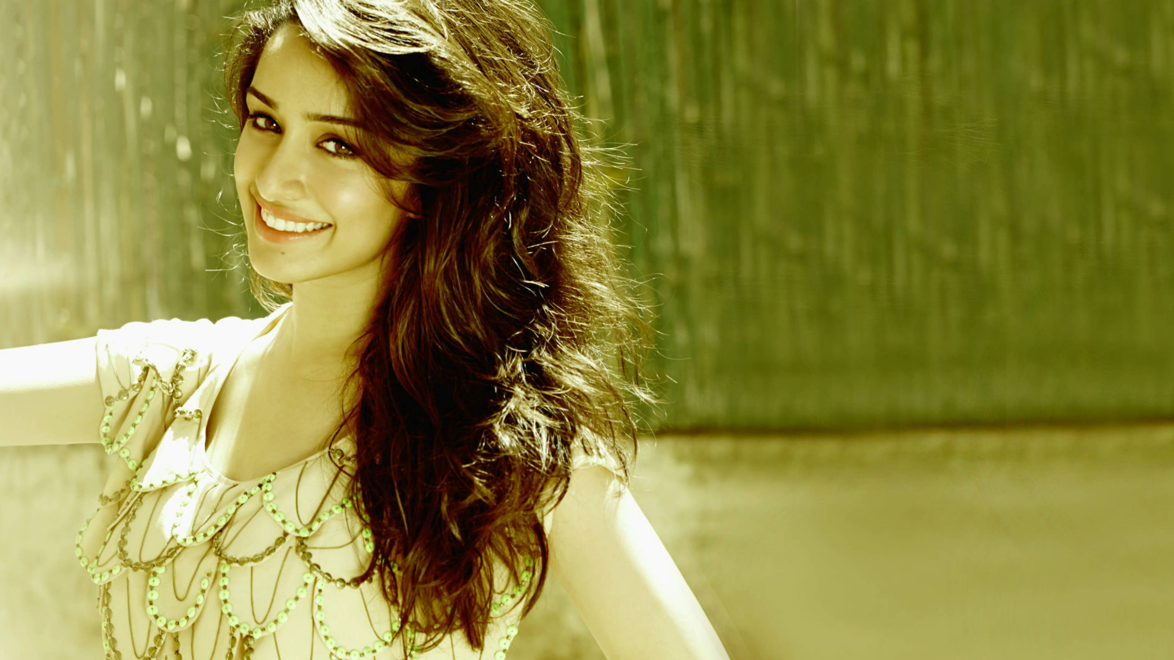 Shraddha Kapoor 桌布 壁紙 — 一位擁有波浪棕色頭髮的燦爛女性，在柔和的金色陽光下，對著有紋理的綠色牆壁溫暖地微笑。