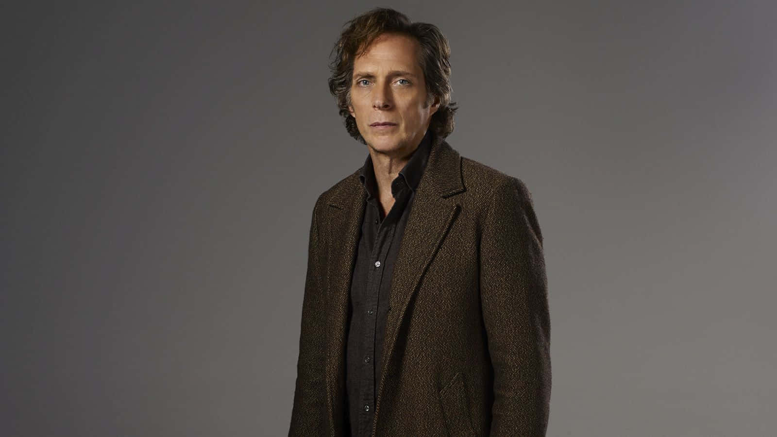 William Fichtner 桌布 壁紙 — 一位擁有波浪棕色頭髮的男子，在純灰色攝影棚背景前，穿著一件質感棕色外套，內搭黑色襯衫。